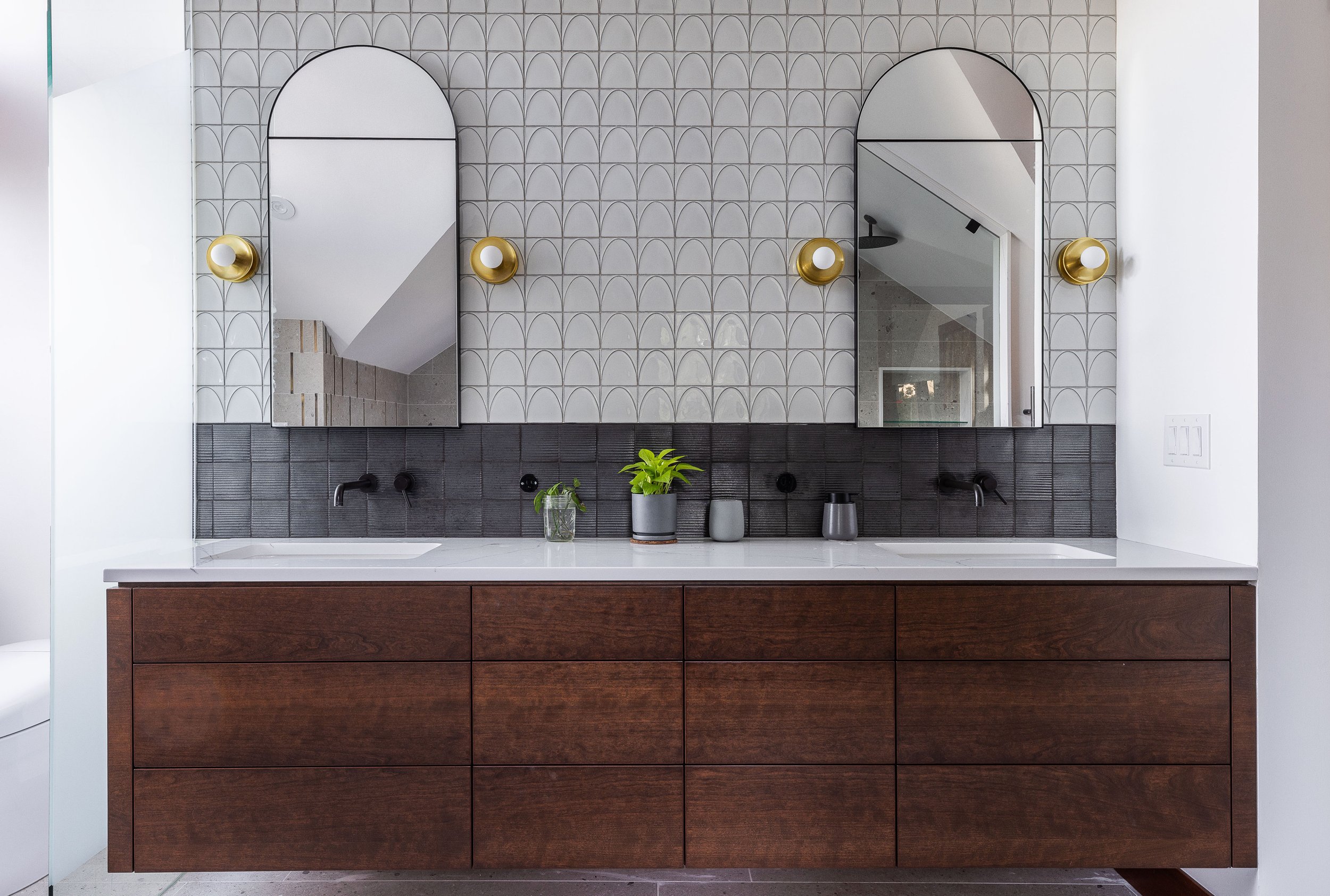 Modern bathroom vanity with double sinks, dark wood cabinets, a white countertop, two mirrors, black wall-mounted faucets, and decorative plants. The wall features white tiles with a scalloped pattern and dark tiles behind the sinks.