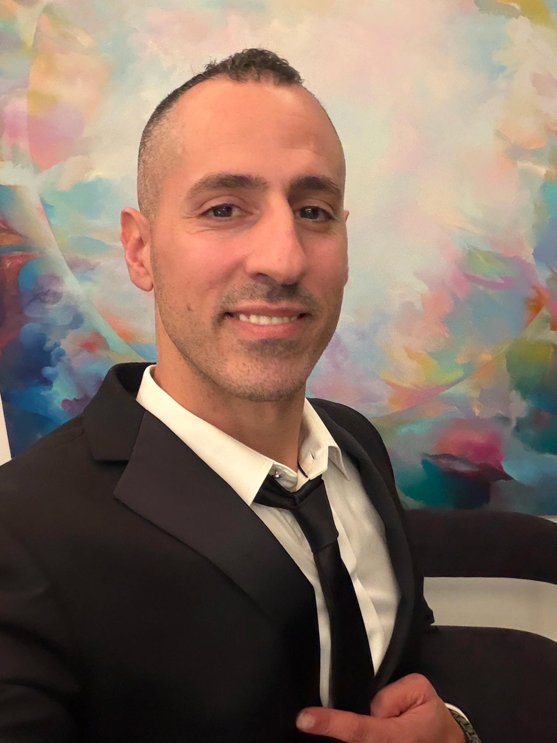 Jonathan Angelilli wearing a tux with a colorful painting behind him. Purestrength.nyc