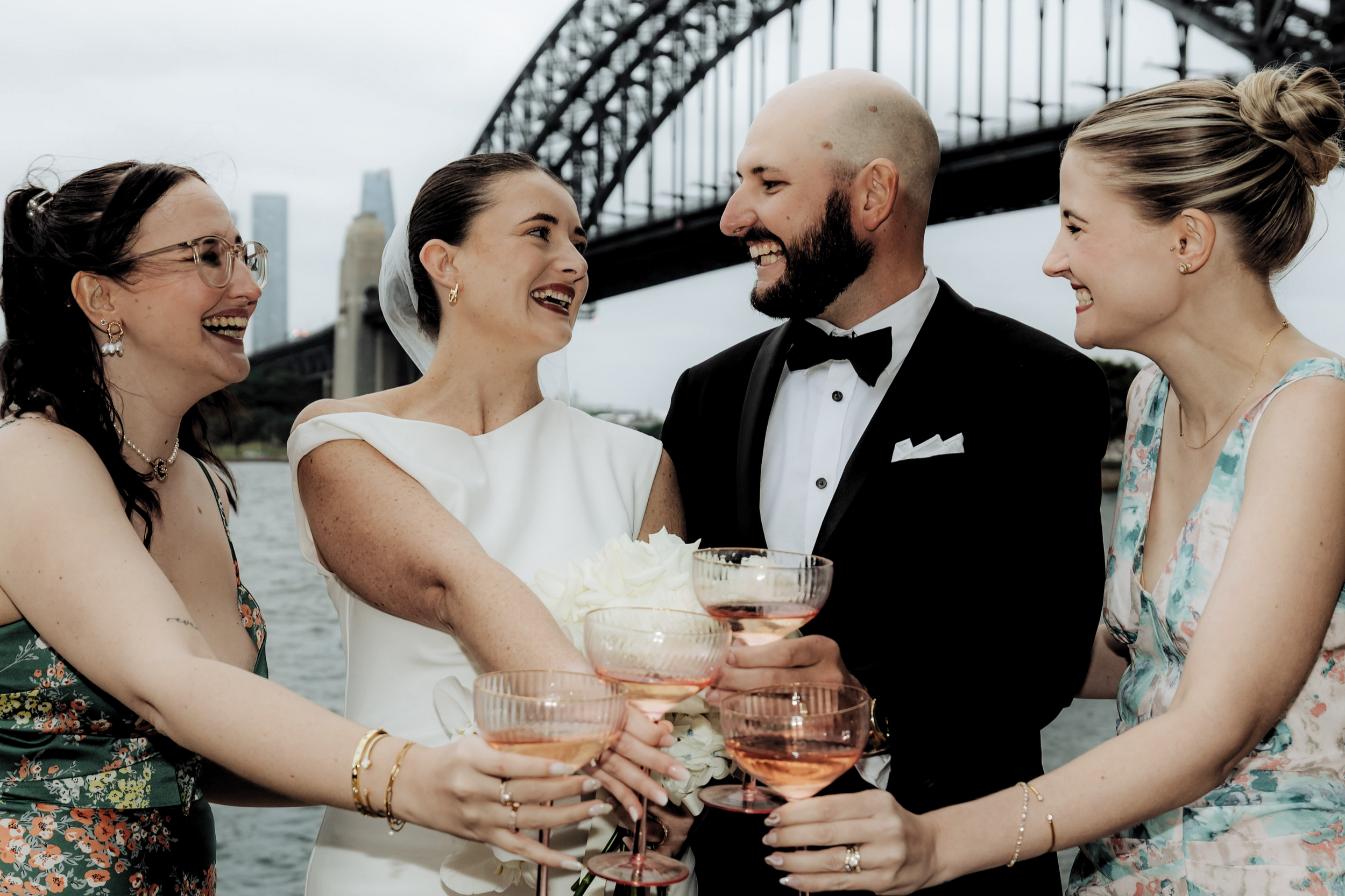 Four people celebrating at a wedding, holding pink drinks and smiling with a bridge and city skyline in the background.