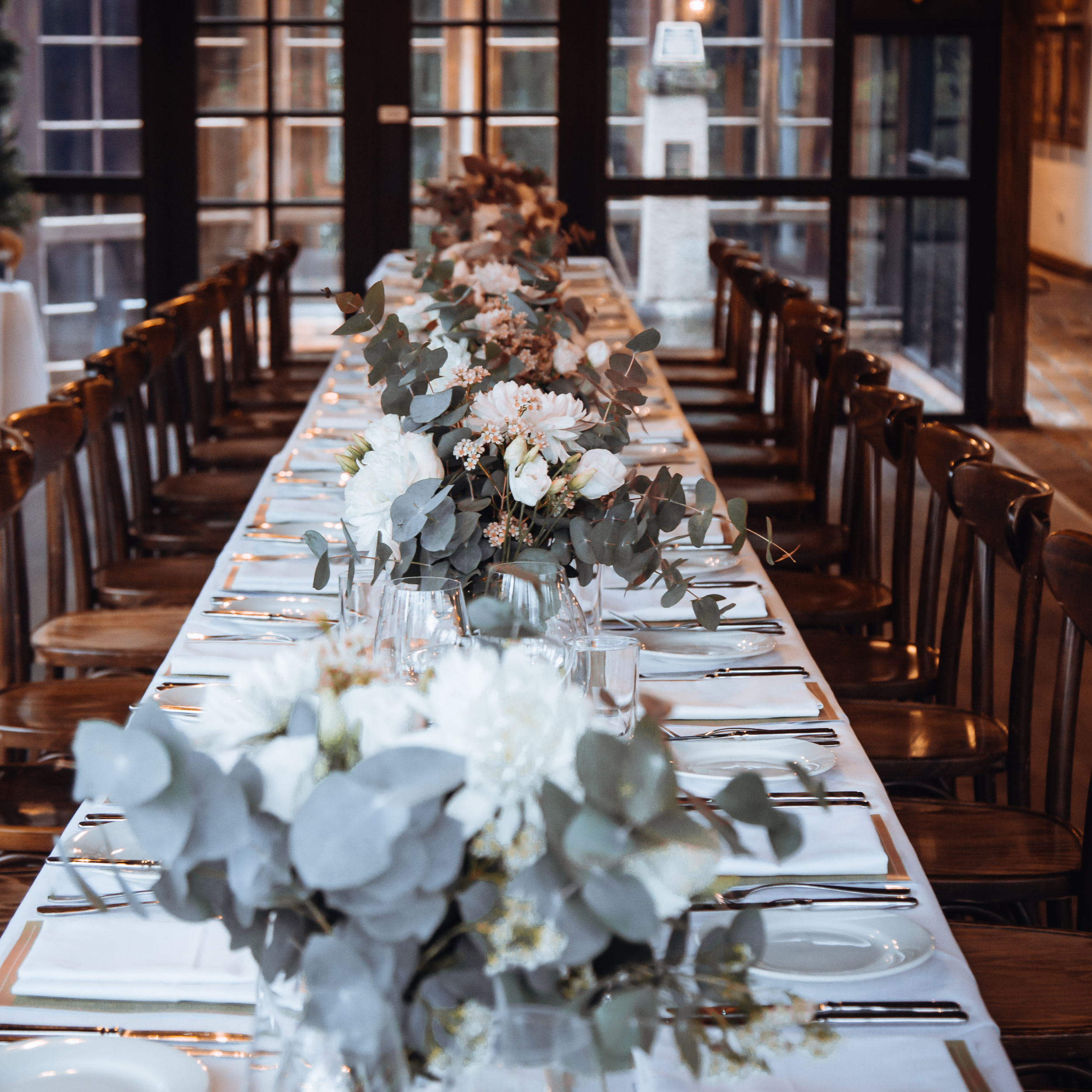 Long banquet table with white tablecloths, decorated with floral centerpieces and place settings, in a well-lit room with large windows.