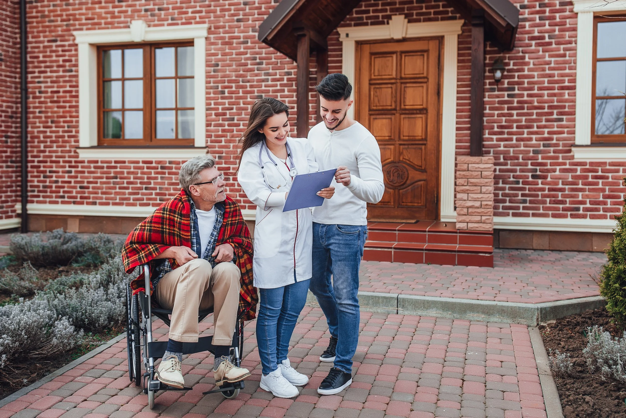 A healthcare professional and a young man stand outside a brick house talking while a senior man in a wheelchair with a blanket over his shoulders watches, smiling.