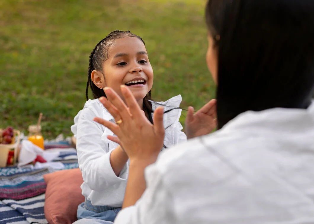A young girl talking and smiling with an adult woman during a picnic outdoors, with a picnic blanket and food in the background.