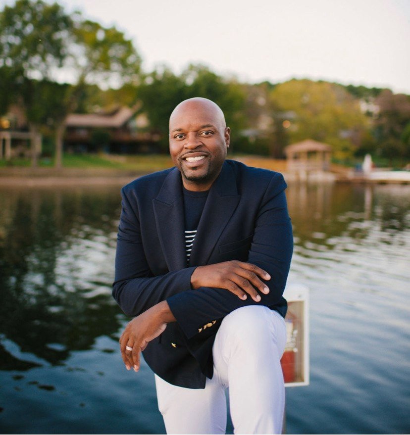 A man sitting by a lake with trees and houses in the background, smiling at the camera, wearing a navy blazer and white pants.
