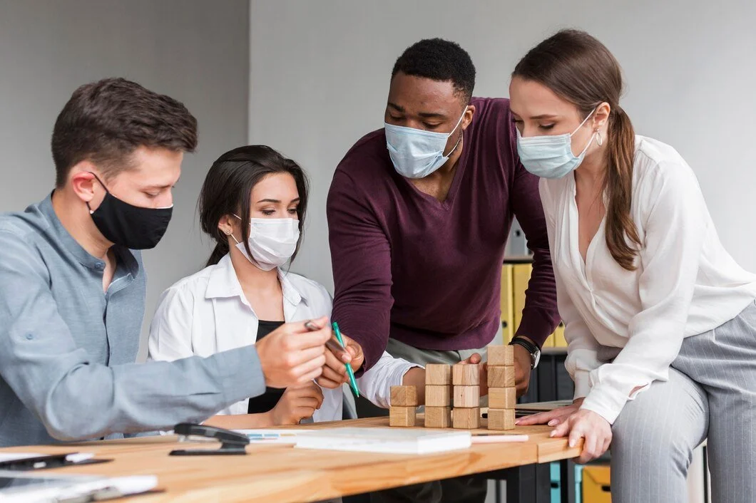 Group of four diverse people wearing face masks working together on a wooden block stacking project at a table in an indoor office or classroom setting.