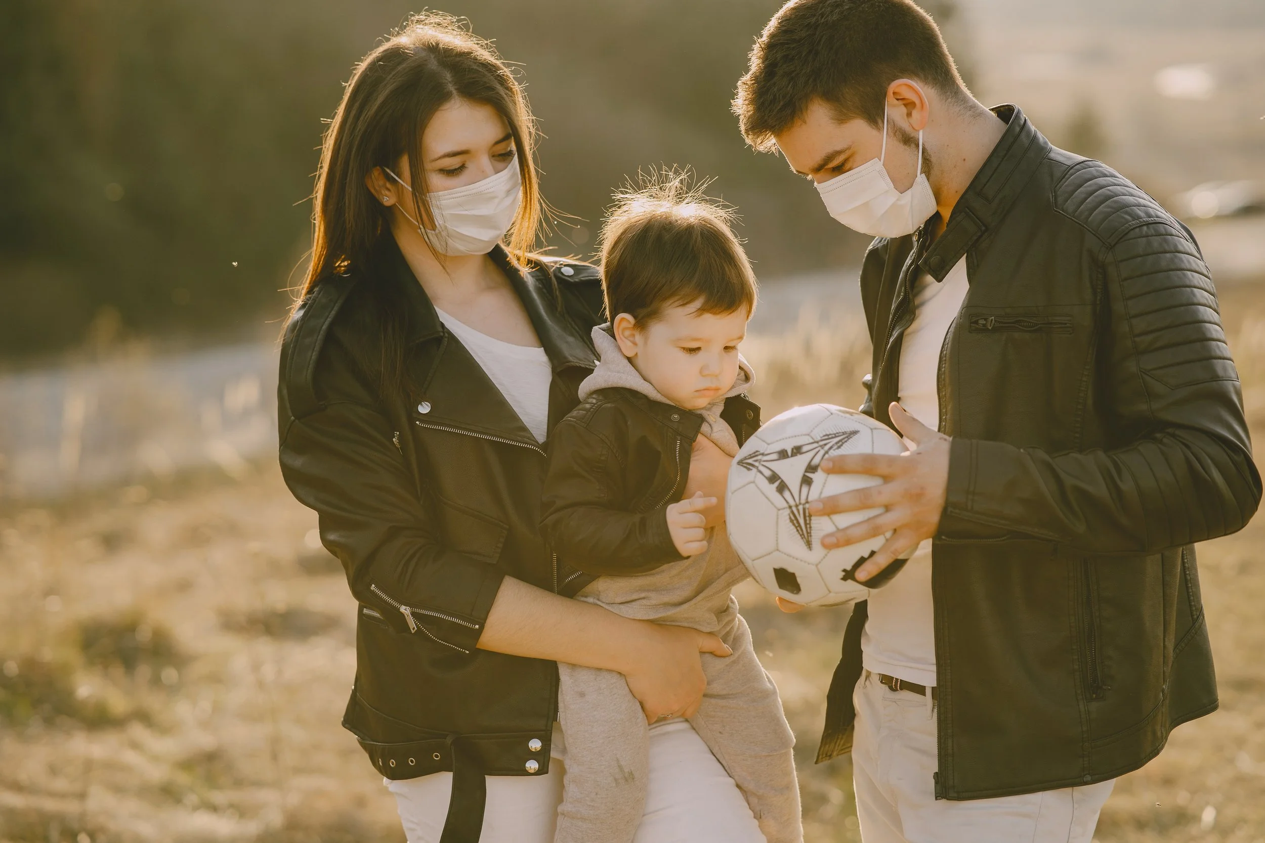 A family of three, wearing masks, stands outdoors in a field. The mother holds a young boy while the father shows him a soccer ball.