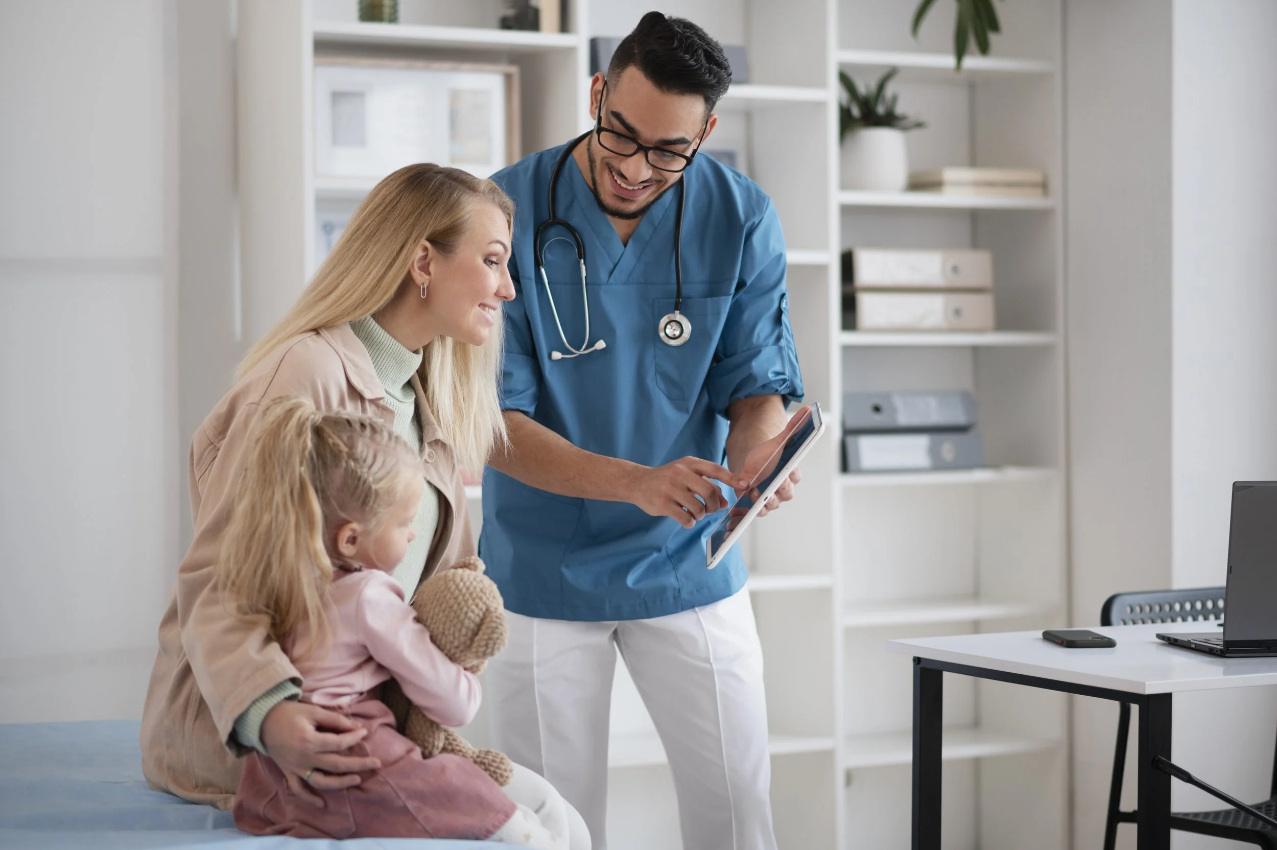 A male healthcare professional in blue scrubs and glasses showing a tablet to a woman with a young girl in a medical office.