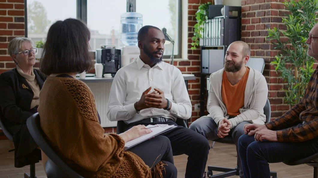 A diverse group of five people engaged in a conversation in a casual meeting room with a brick wall background, plants, and large windows.