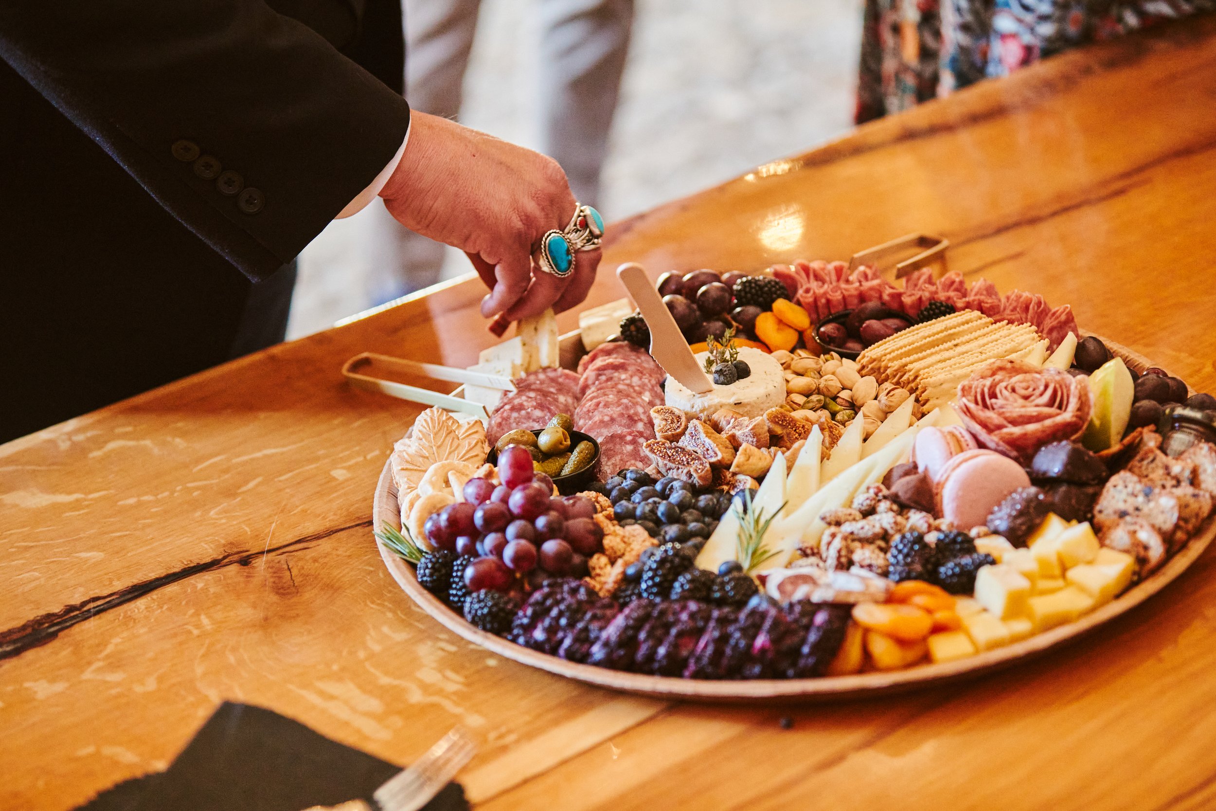 A person wearing a black suit with gray buttons is reaching towards a large cheese and charcuterie board filled with various cheeses, grapes, berries, nuts, crackers, and sliced meats on a wooden table.