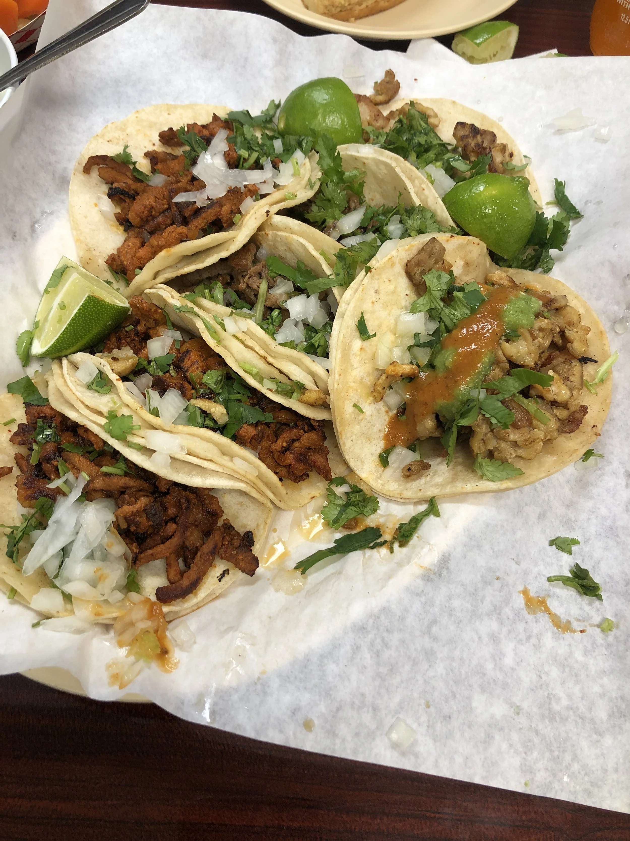 Three tacos with grilled meat, shredded cheese, onions, cilantro, and lime wedges on a white paper-lined tray.
