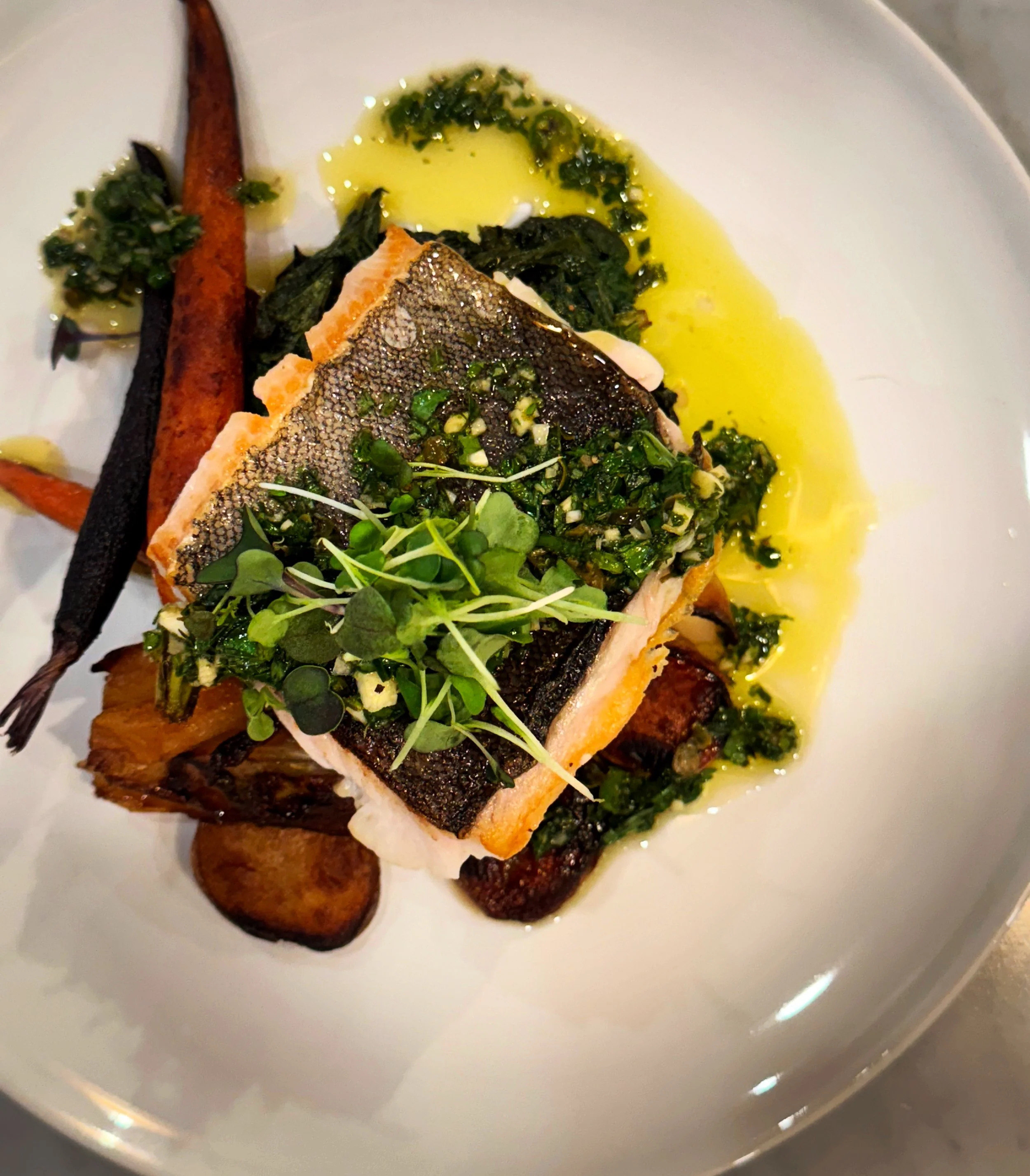 A plated dish featuring a fillet of cooked fish with crispy skin, topped with chopped herbs, served on a bed of roasted vegetables, and garnished with microgreens, with a drizzle of yellow sauce or oil.