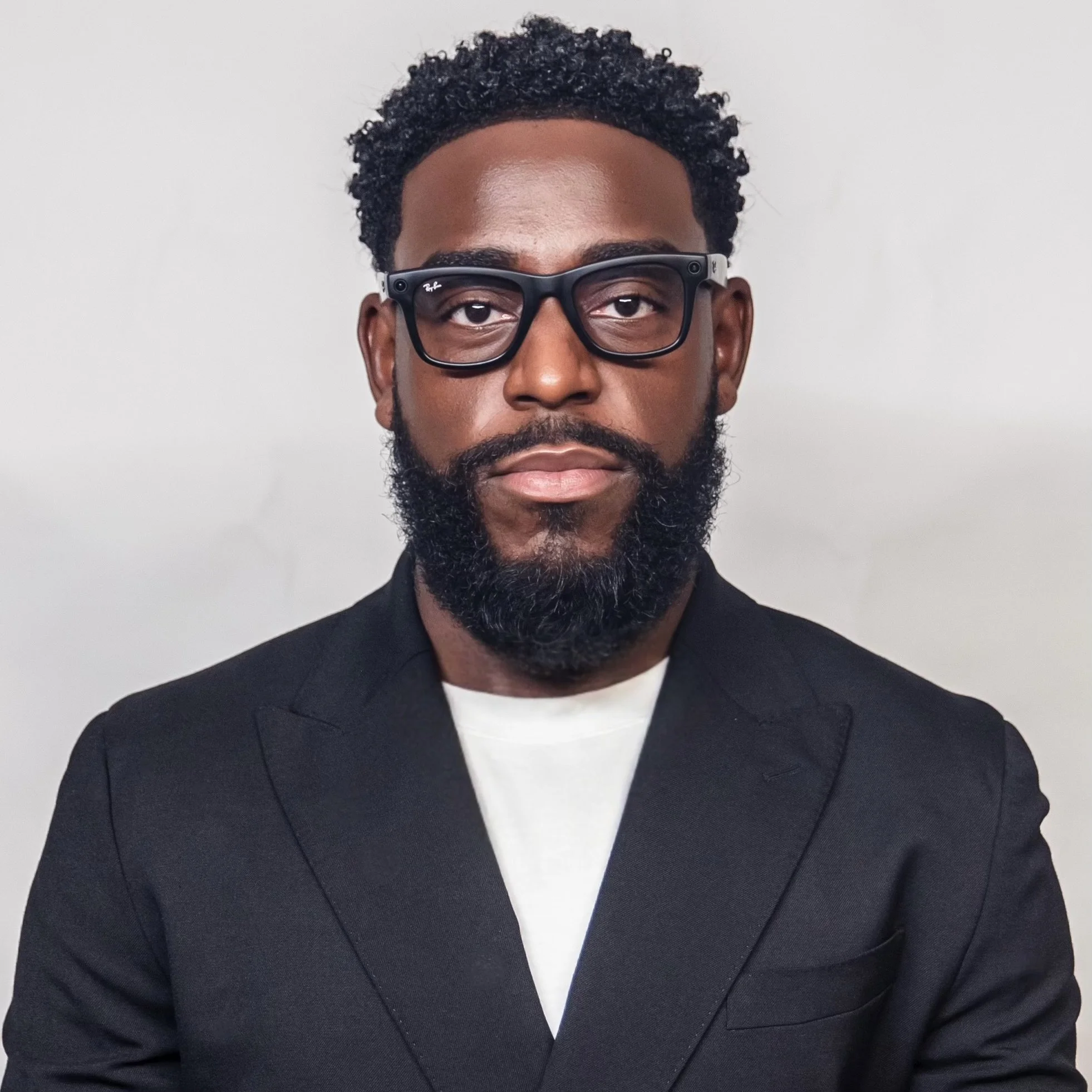 A man with dark skin, curly hair, and a beard wearing black glasses and a black blazer over a white shirt, posing against a plain white background.