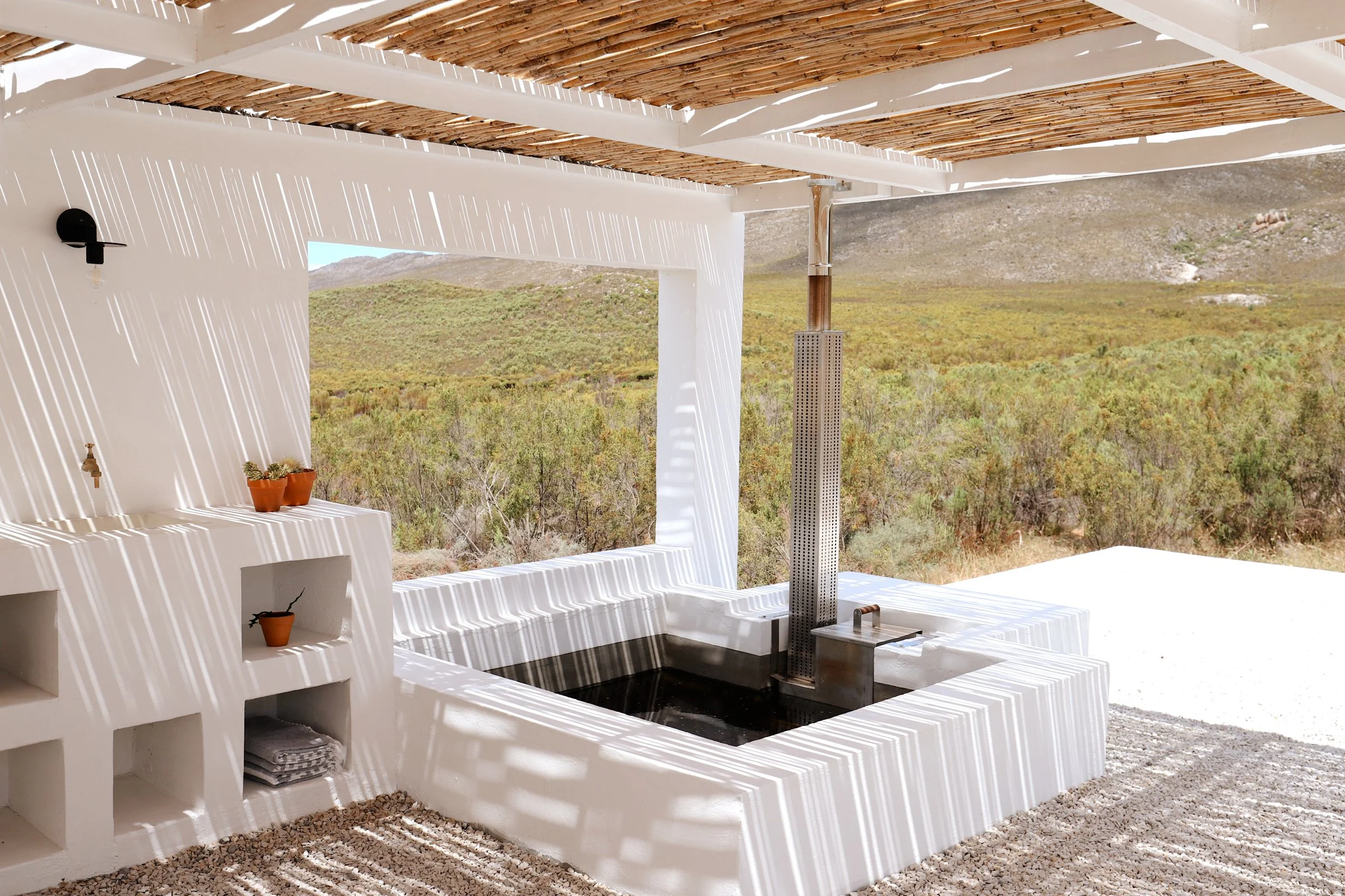 Modern outdoor spa with white plaster seating, bamboo roof, and surrounding nature.