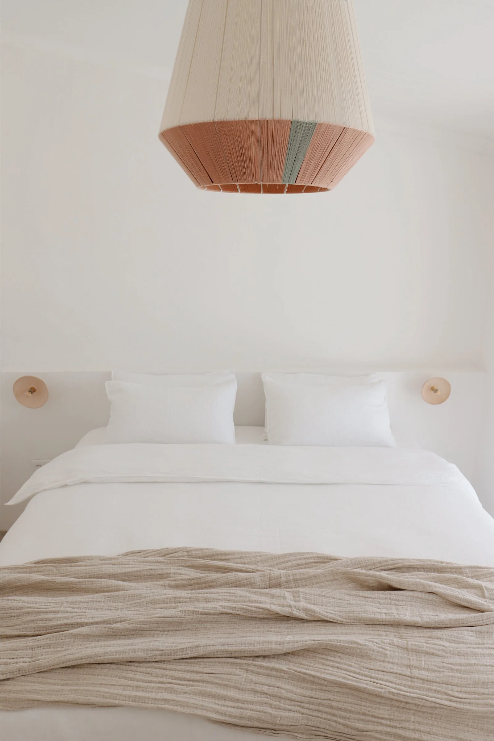 Minimalist bedroom with white bedding, light brown textured throw, and a large woven pendant lamp above.