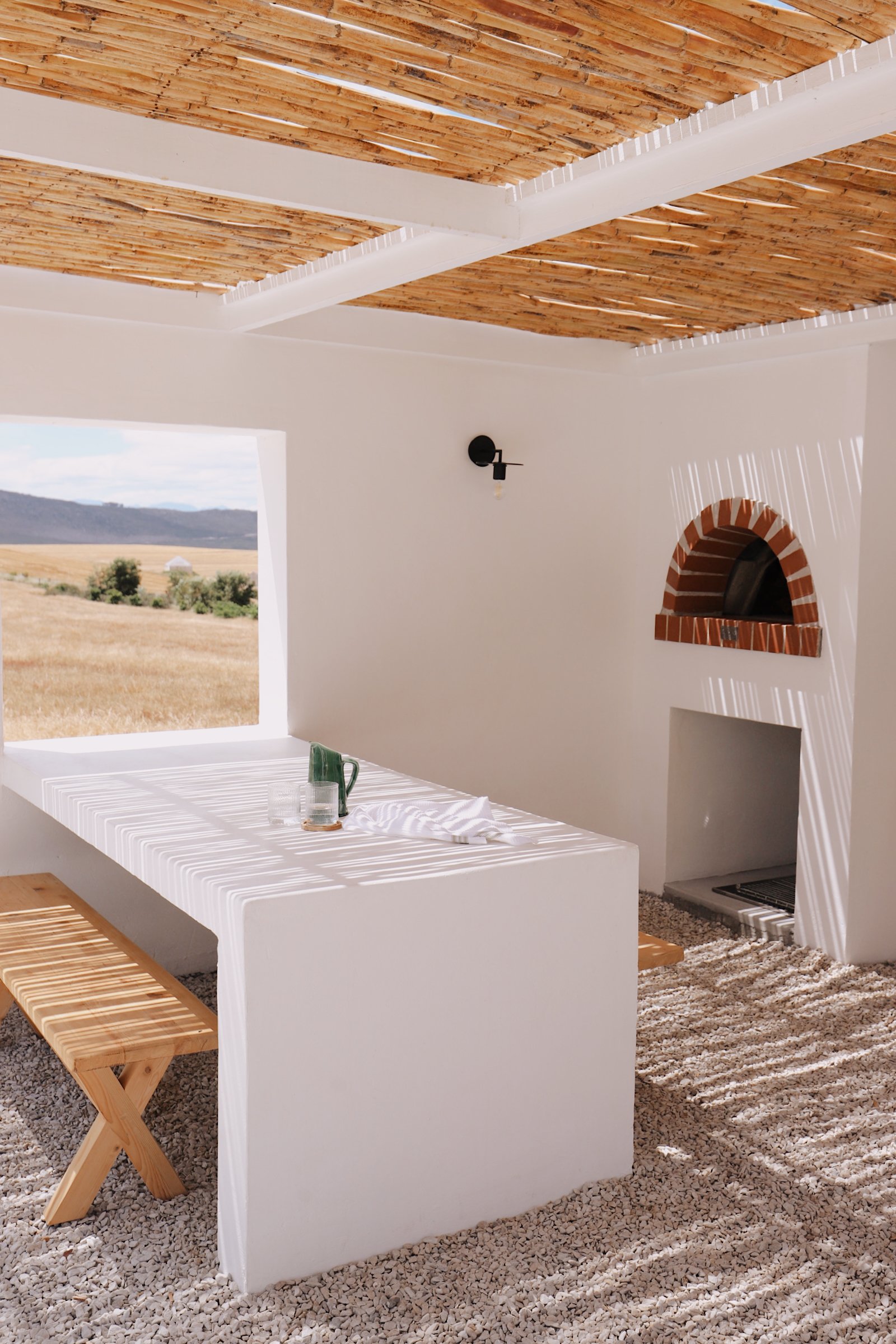 White outdoor dining area with a rectangular table and bench, under a bamboo pergola. Walls are white with a small arched pizza oven and a window opening to a landscape view. Pebbles cover the ground.