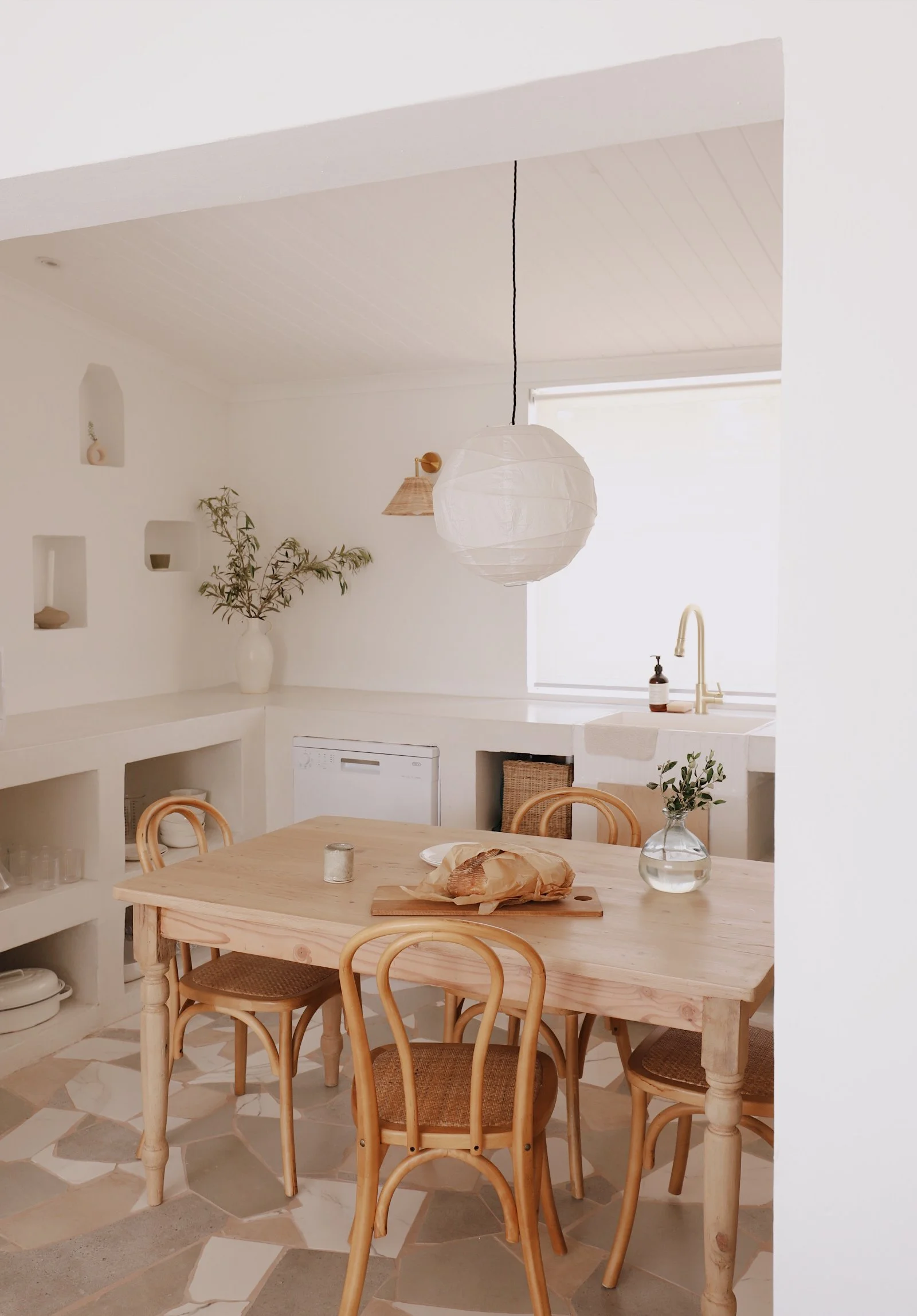 Minimalist kitchen with a wooden dining table and chairs, pendant lamp, white walls, and natural décor. A loaf of bread is on the table, with potted greenery nearby.