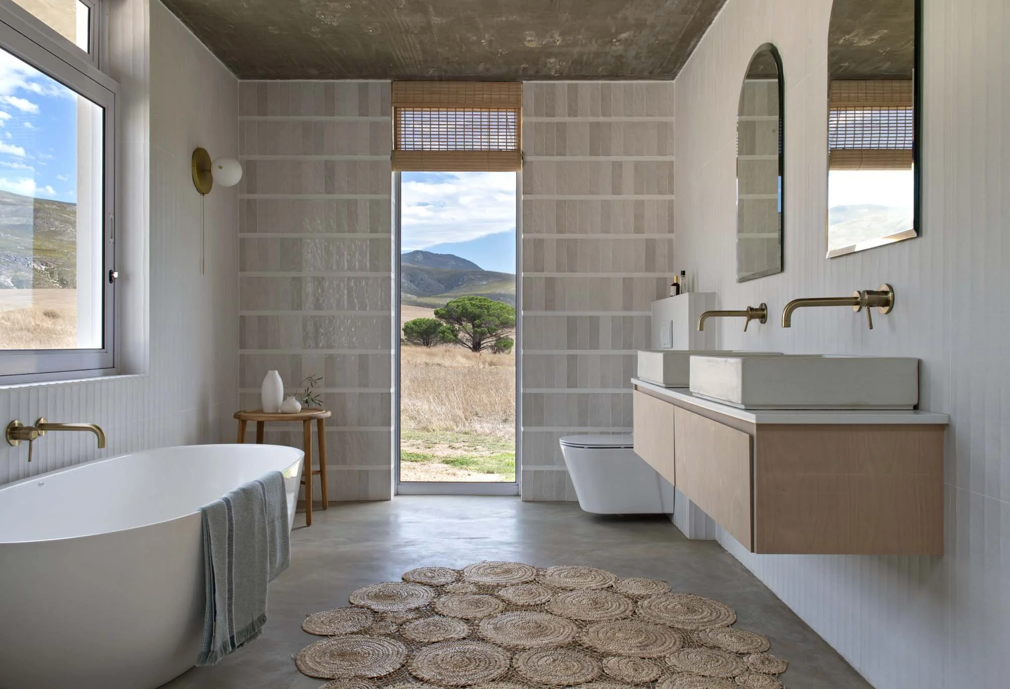 Modern bathroom with freestanding bathtub, double sink vanity, large mirrors, and textured wall. A window reveals an outdoor landscape with hills and a tree. Light enters through the window and door.