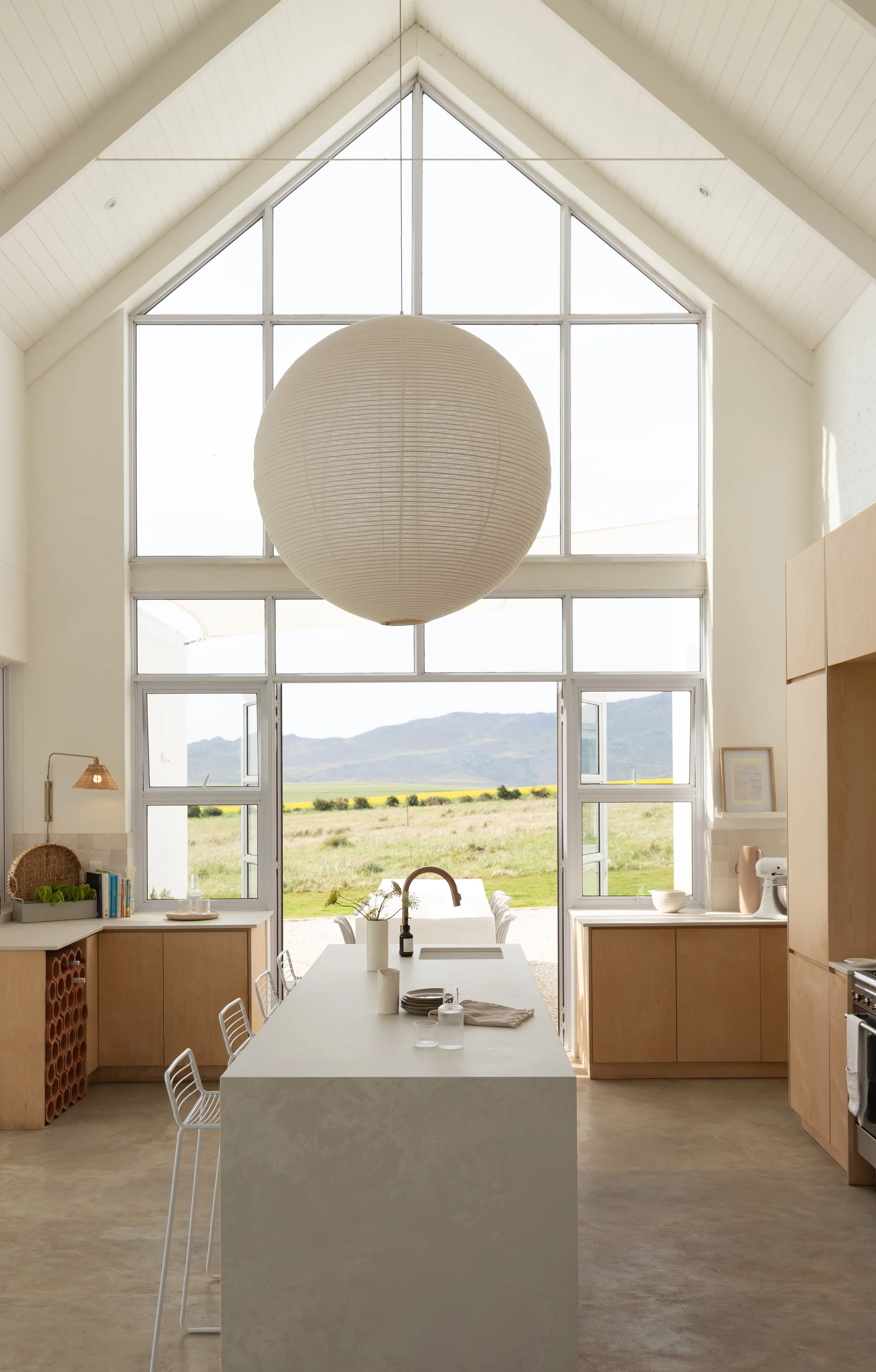Modern kitchen with high ceiling, large windows, wooden cabinetry, a central island with white countertop, metal bar stools, a wicker pendant light, and a view of an open landscape.