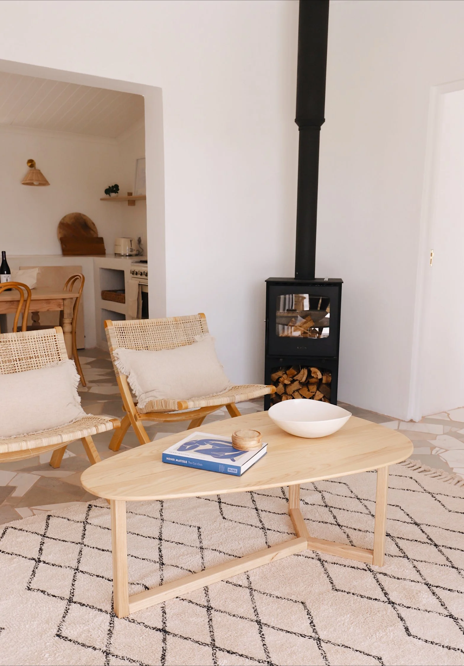 Cozy living room with a wood-burning stove, wicker chairs, a wooden coffee table with a book and bowl, and a patterned rug. Adjacent kitchen area with wooden furniture and light décor.