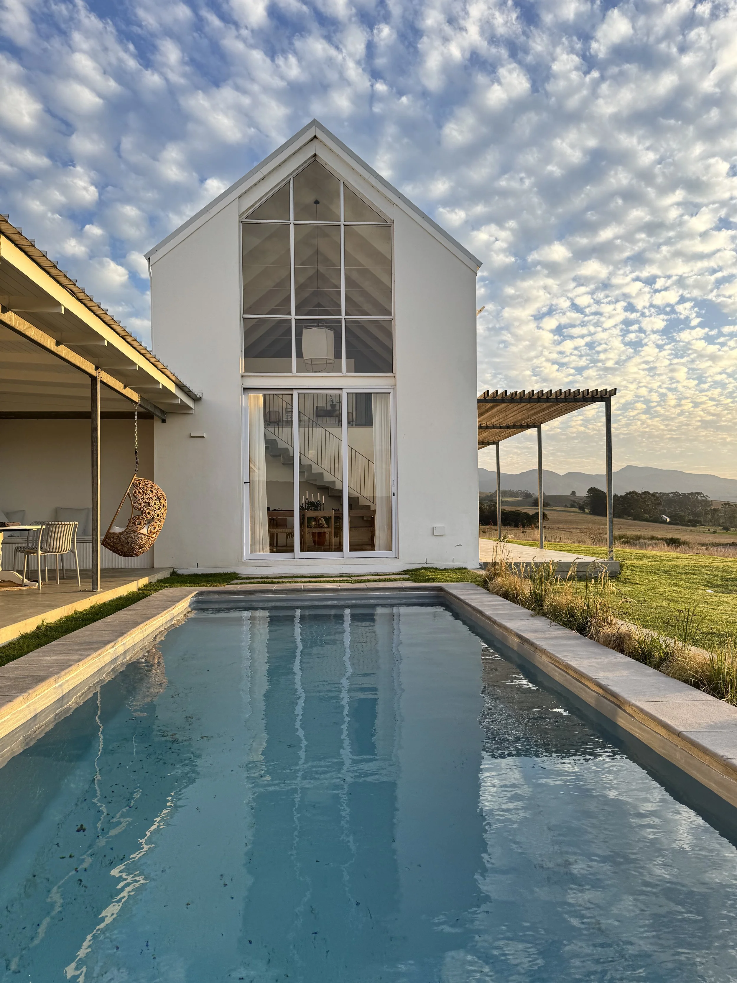 Modern open-plan interior with large windows showcasing a view of mountains and a river. Features a wicker light fixture, dining table with wooden chairs, a minimalist kitchen island, and a decorative plant.