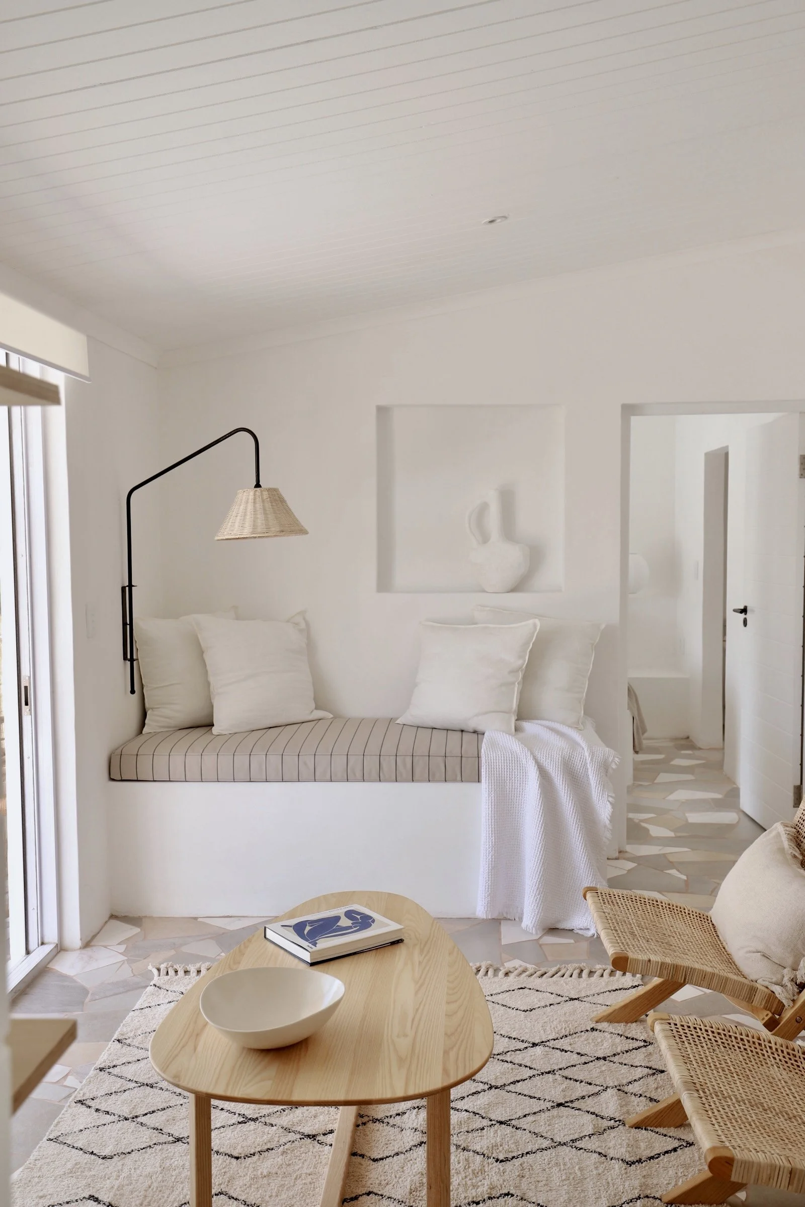 Minimalist living room with white walls, a built-in bench with striped cushion and white pillows, a wall-mounted lamp, wooden coffee table with a bowl and book, woven chairs, and a cream rug.