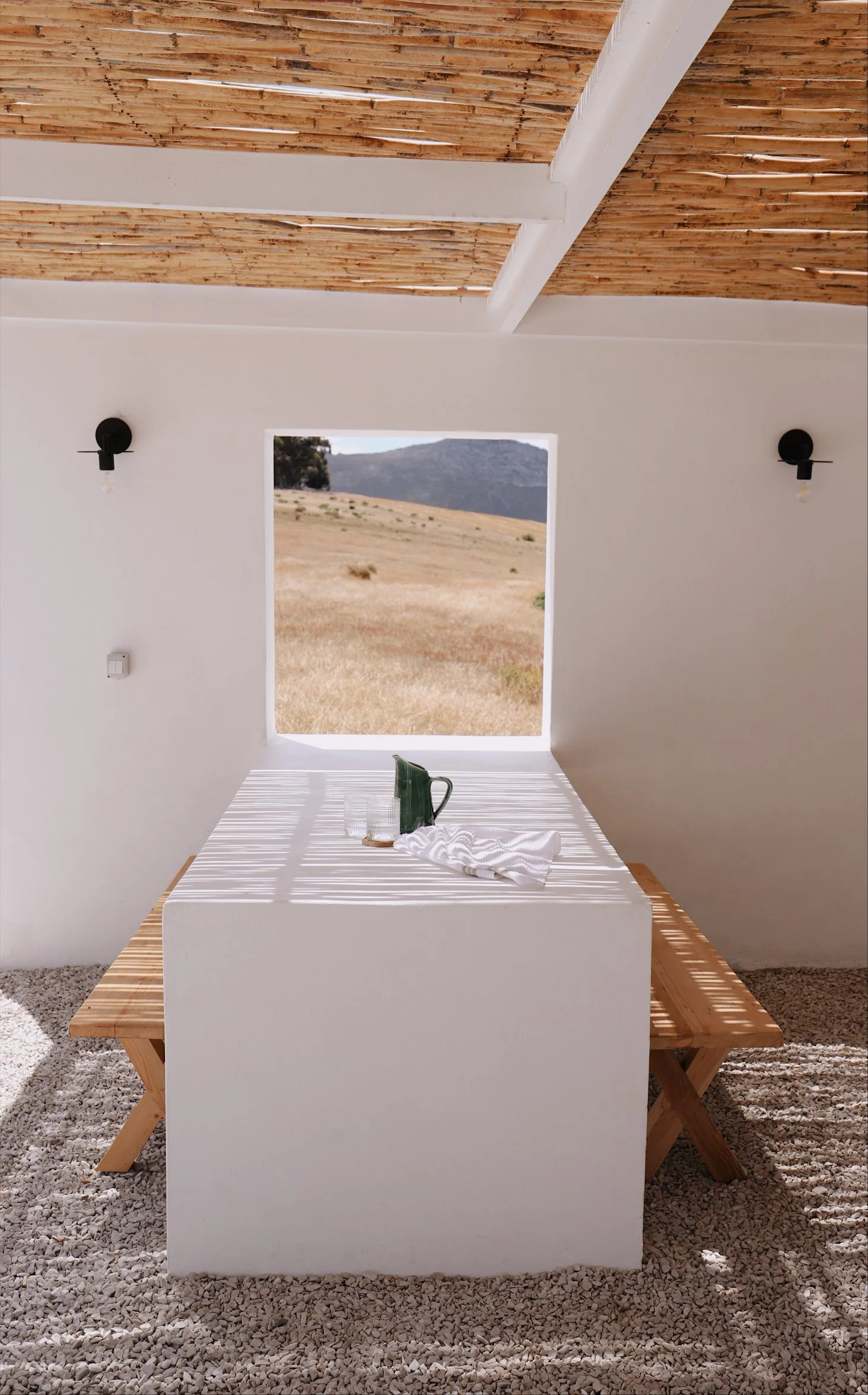 Outdoor dining area with a minimalist table and benches under a pergola. Pebble ground, white walls, wooden ceiling, and scenic landscape view through a window.