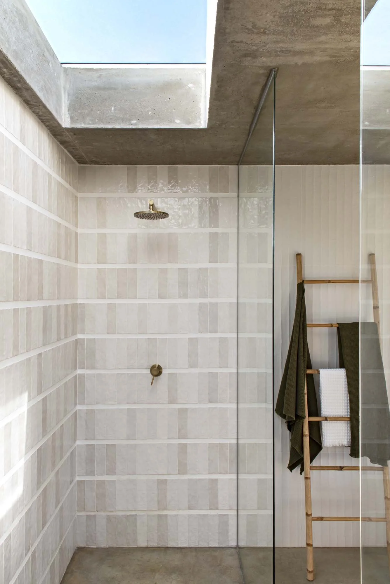 Minimalist bathroom shower with beige tiled walls, skylight, rain shower head, glass partition, and bamboo ladder with towels.