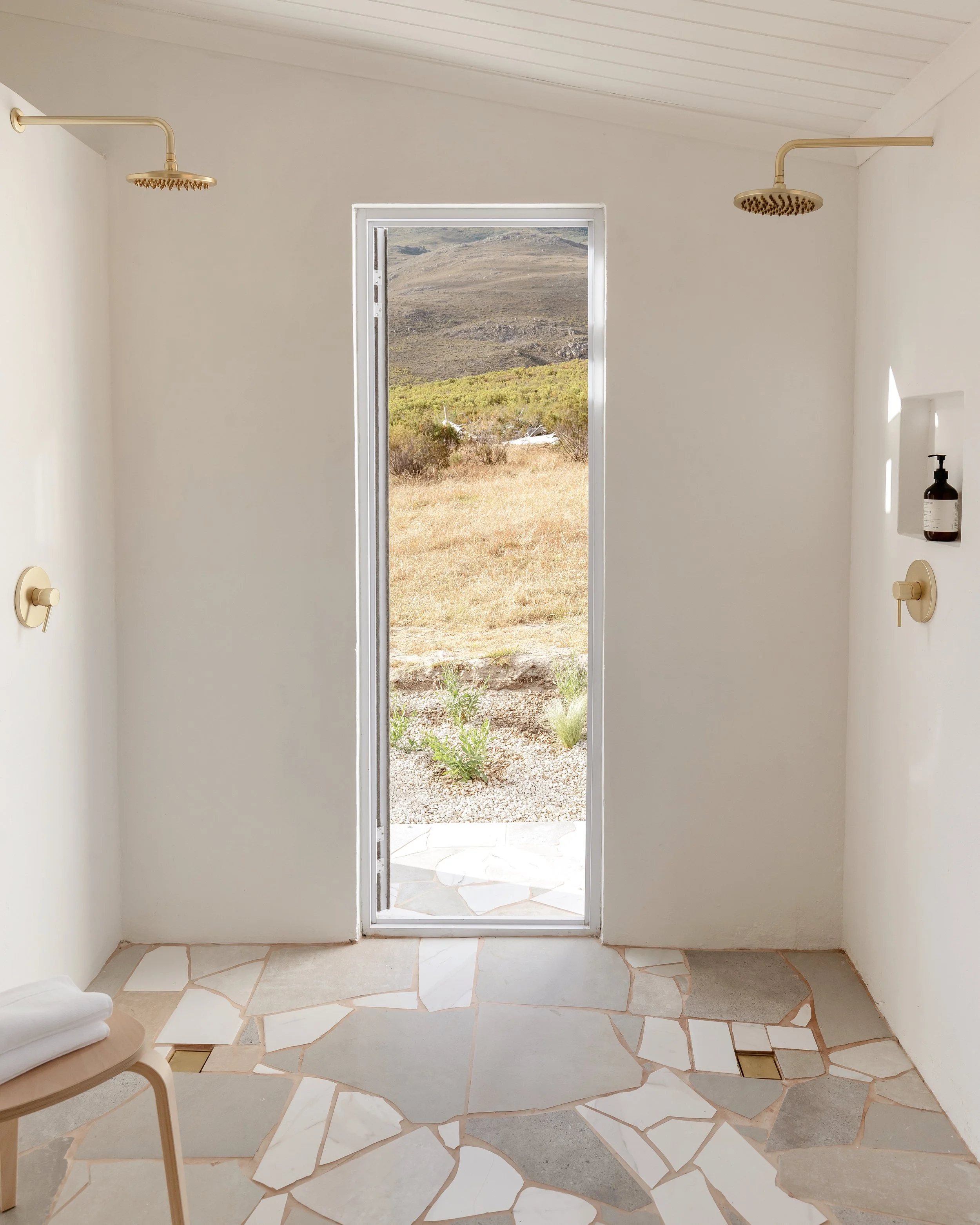 Minimalist bathroom with two shower heads and a door opening to an outdoor landscape view. Features light stone flooring, a small side table, and wall-mounted soap dispenser.
