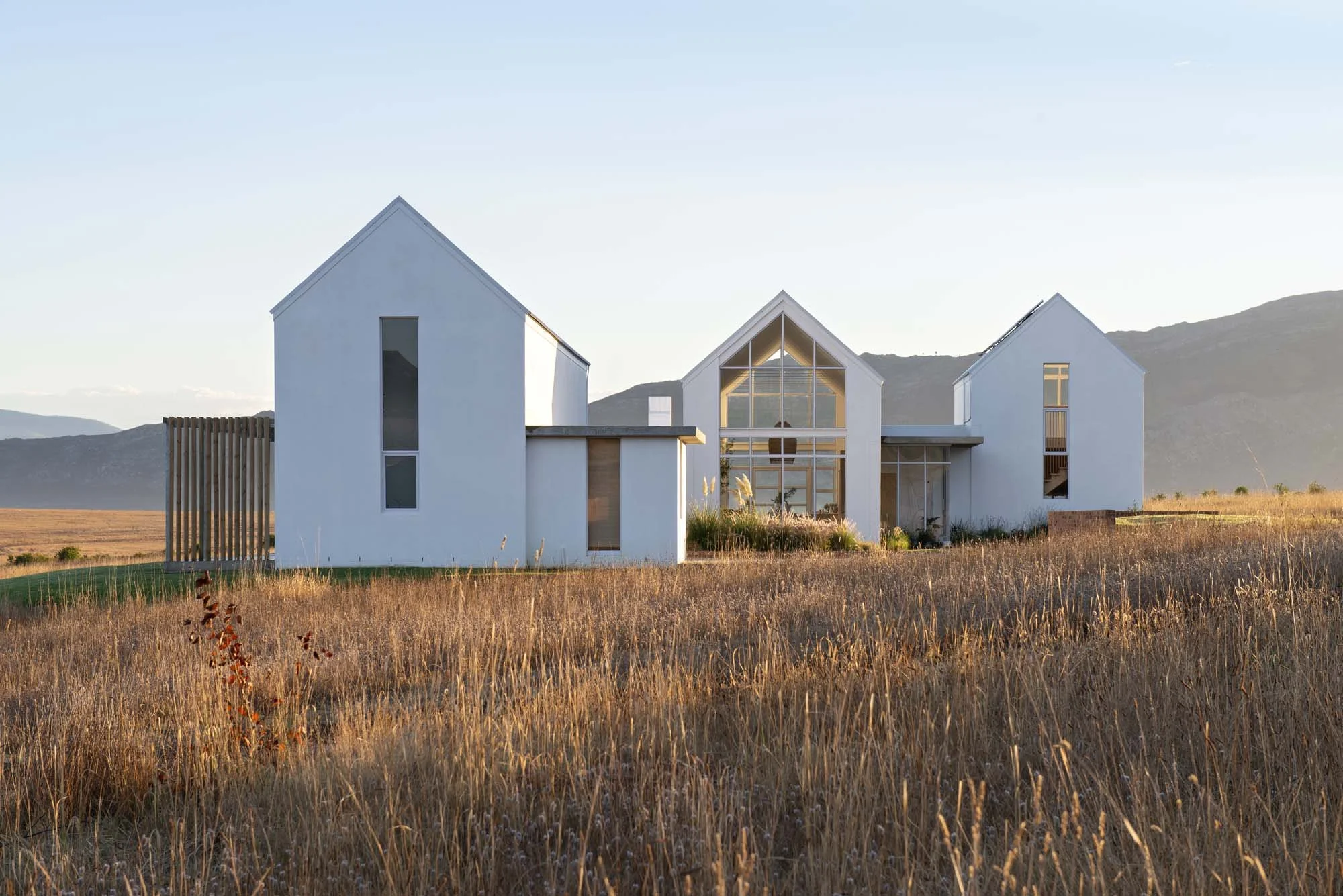 Modern rural house with white exterior and large windows, surrounded by dry grassland and mountains in the background.