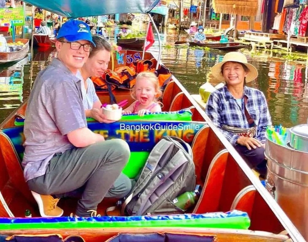 A family of four enjoying a boat ride on a canal at a floating market in Thailand, with colorful market stalls in the background.