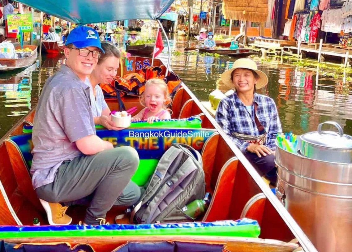 People in a boat on a bustling floating market in Thailand, with colorful market stalls, boats, and vendors in the background.