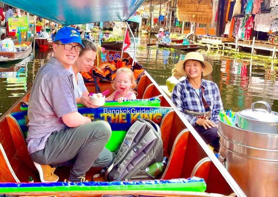 A group of four people enjoying a boat ride through a floating market in Bangkok, Thailand. They are sitting in a traditional longtail boat surrounded by colorful market stalls, water, and vendors.