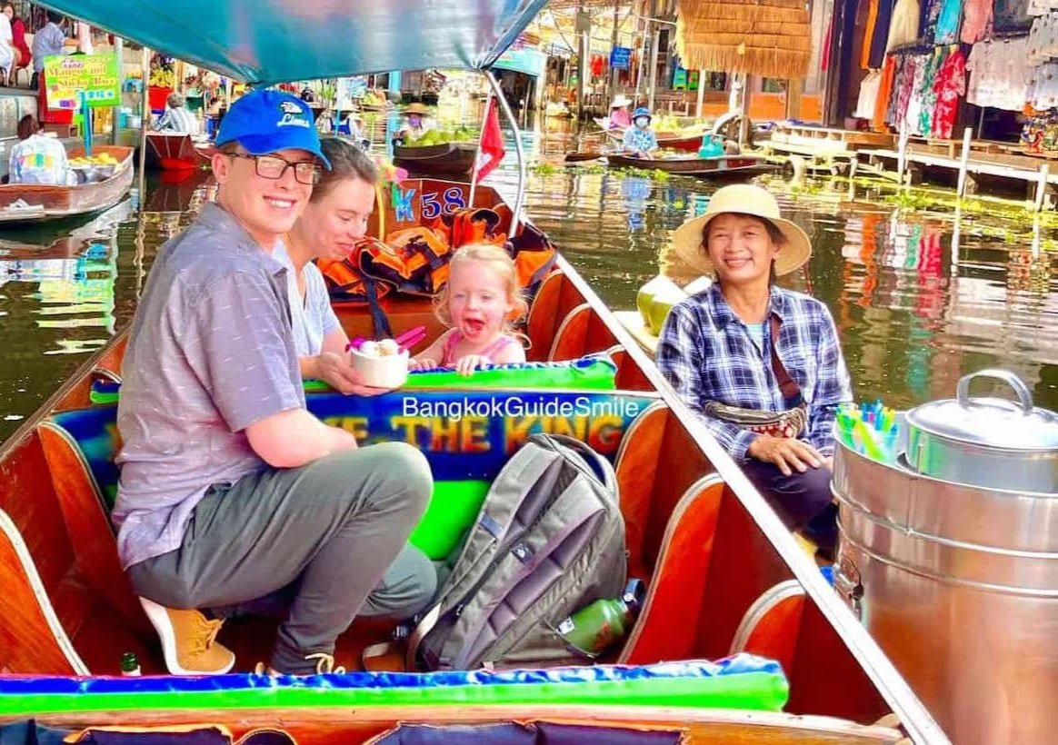 People on a boat tour along a floating market in Bangkok, Thailand. The group includes a young man, a woman, a young girl, and a local boat operator. The scene is vibrant with colorful market stalls in the background, and water reflections on the canal.