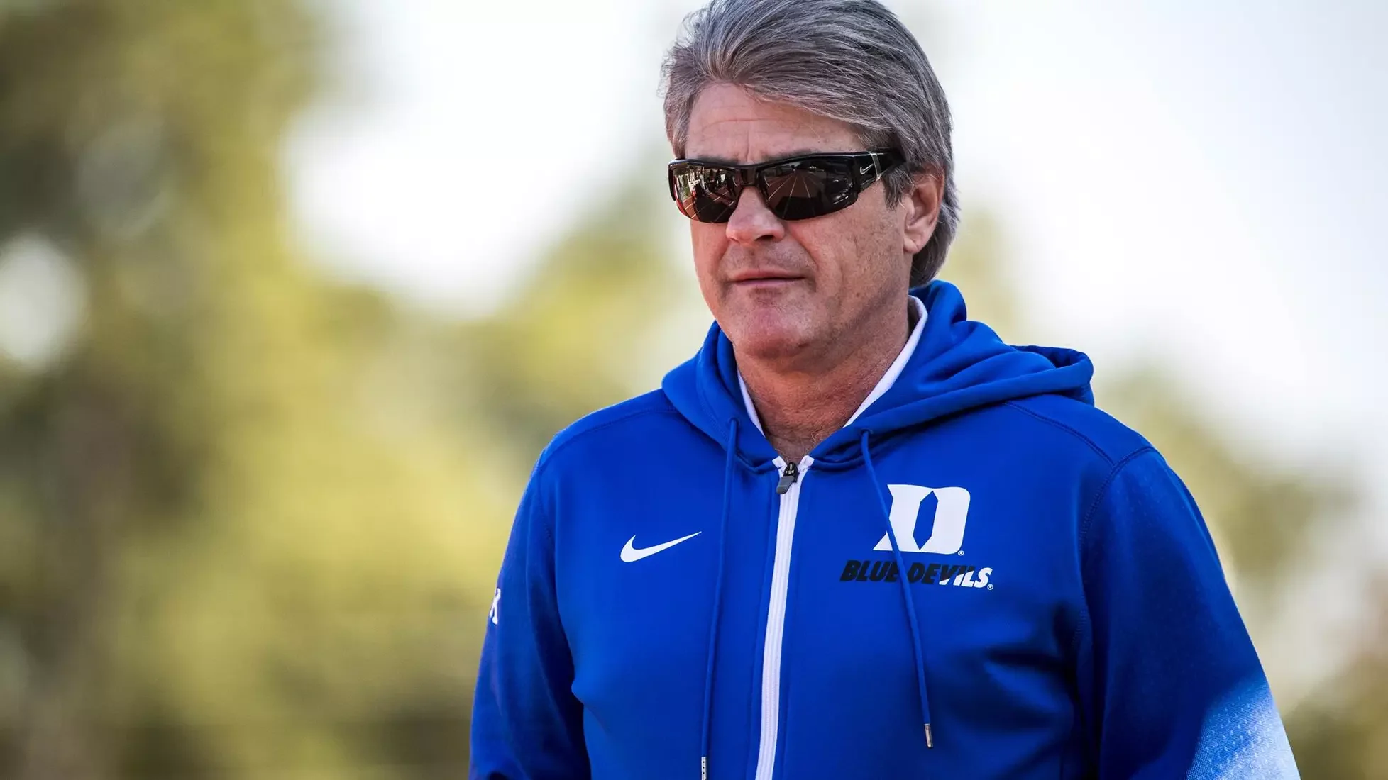 A middle-aged man with gray hair wearing sunglasses and a blue hoodie with a Nike logo and 'Blue Devils' text.