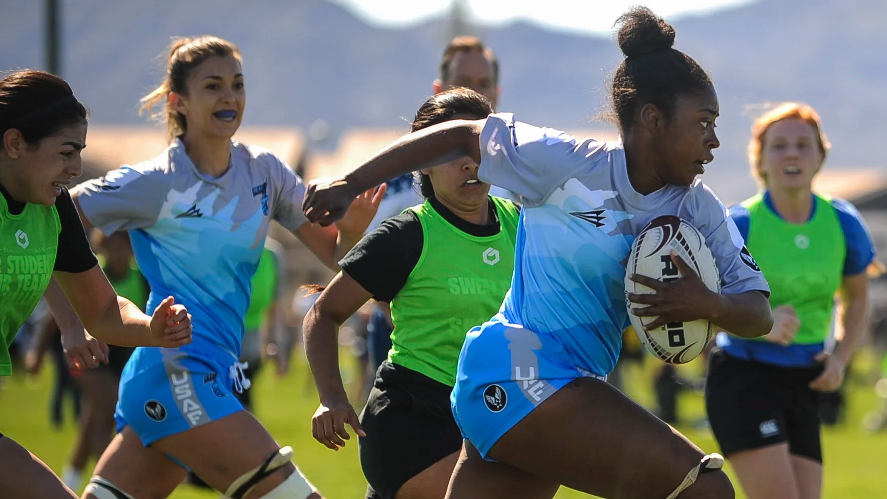 Women playing rugby on a field, with one woman carrying the ball and others running nearby, wearing sports uniforms.