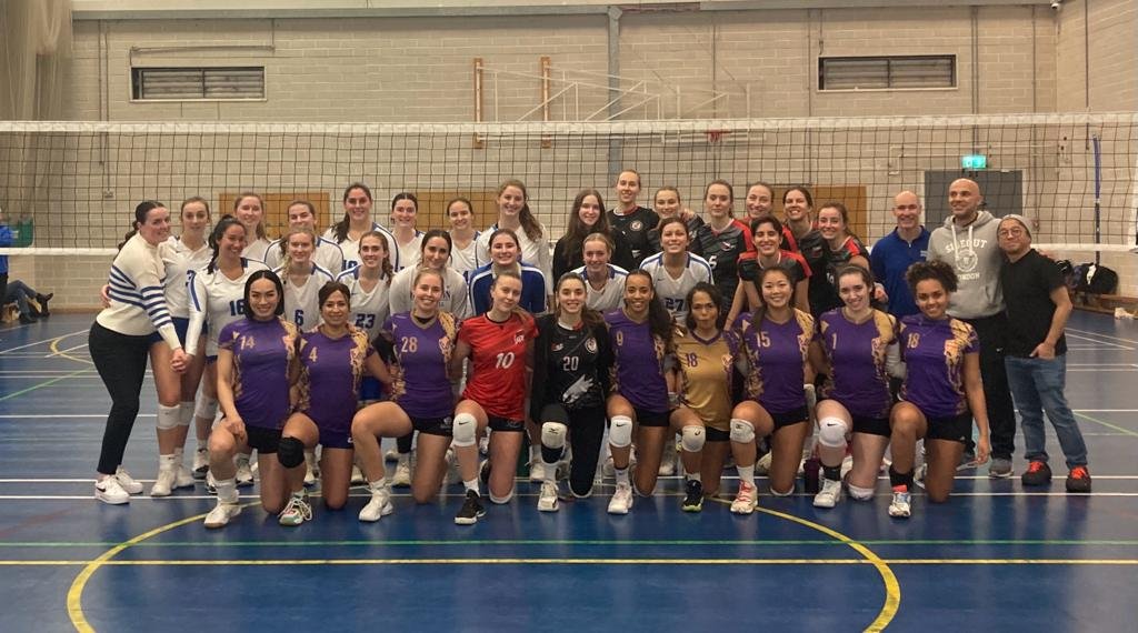 Group of female volleyball players posing in a gymnasium, with spectators and coaches in the background.