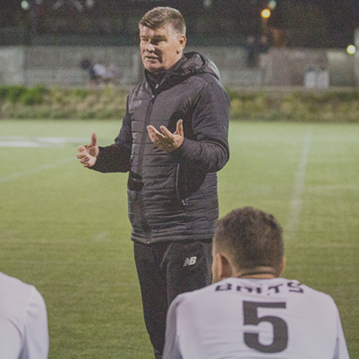 A coach in a black puffer jacket giving instructions to a football team on the sideline during a game at night.