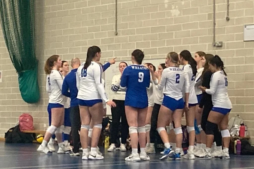 A group of female volleyball players in white and blue uniforms huddled together during a game, on an indoor court with a brick wall background.