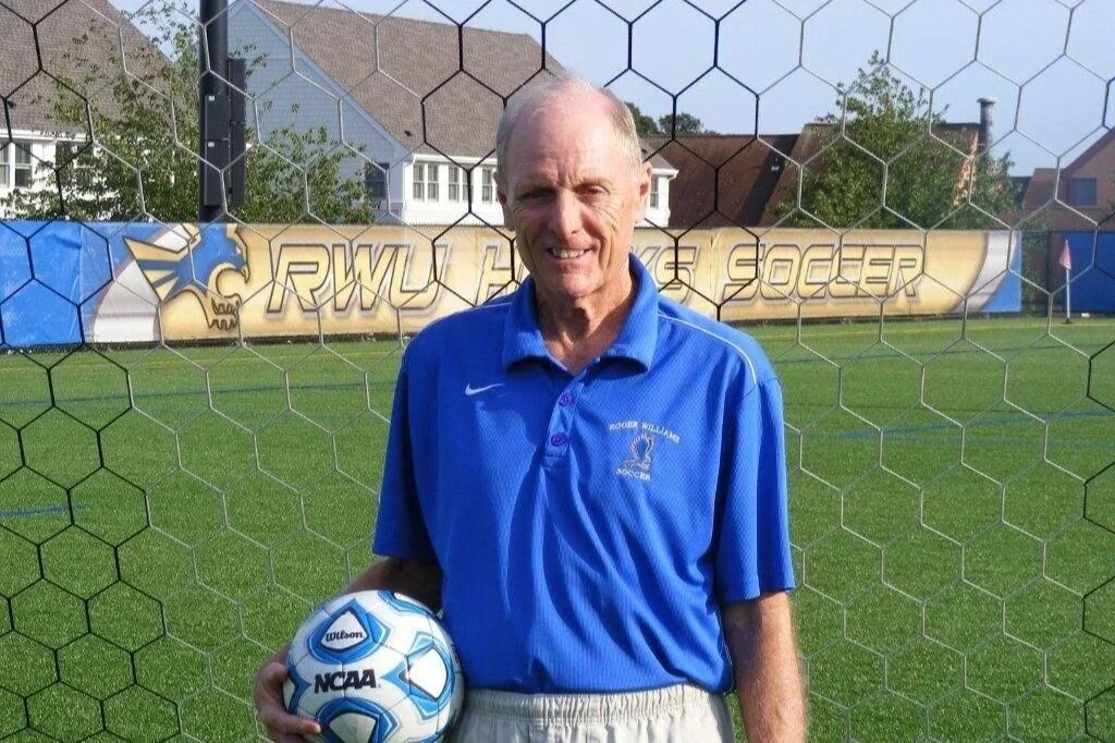 An elderly man standing on a soccer field behind a goal net, holding a soccer ball, smiling at the camera. He is wearing a blue sports polo shirt and beige shorts, with a soccer-related logo on his shirt. In the background, there are houses, trees, and a banner that reads 'RWU Tigers Soccer.'