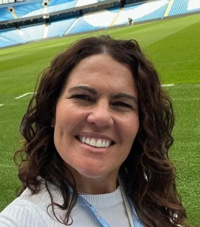 A woman with long, curly brown hair smiling at the camera in a stadium with green grass and empty blue and yellow seats.