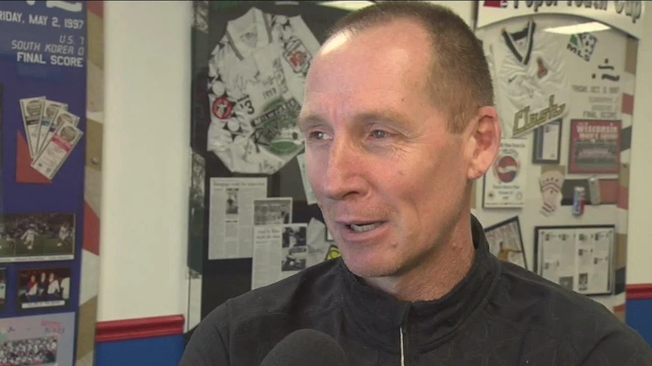 A man being interviewed, with sports memorabilia and newspaper clippings in the background, in a sports setting.