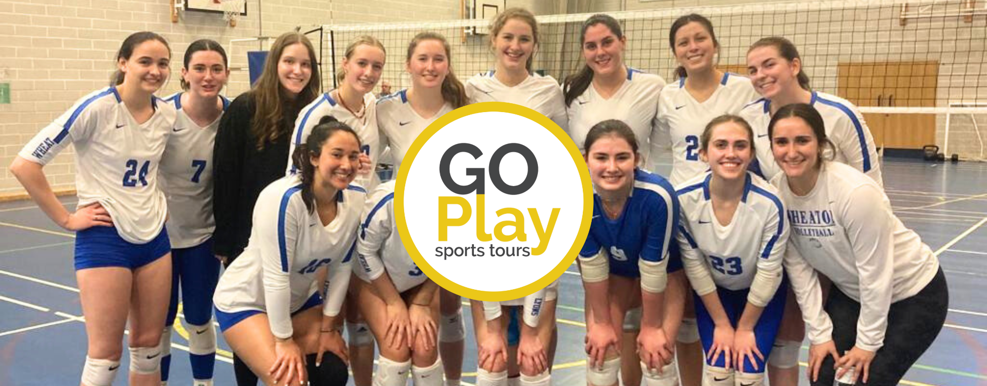 A group of eleven young women volleyball players in white and blue uniforms poses together in a gymnasium. They are smiling, with some kneeling in front and others standing behind them. The gym features a volleyball net and court markings. Overlaid at the center is a circular logo with the text 'GO Play sports tours.'