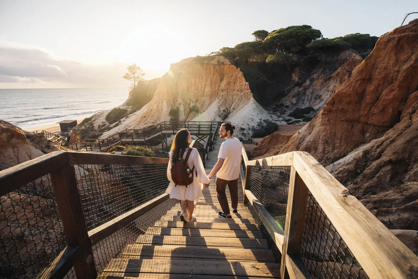 Un couple marche main dans la main sur une passerelle en bois au bord de la mer, entouré de falaises avec des arbres, au coucher du soleil.