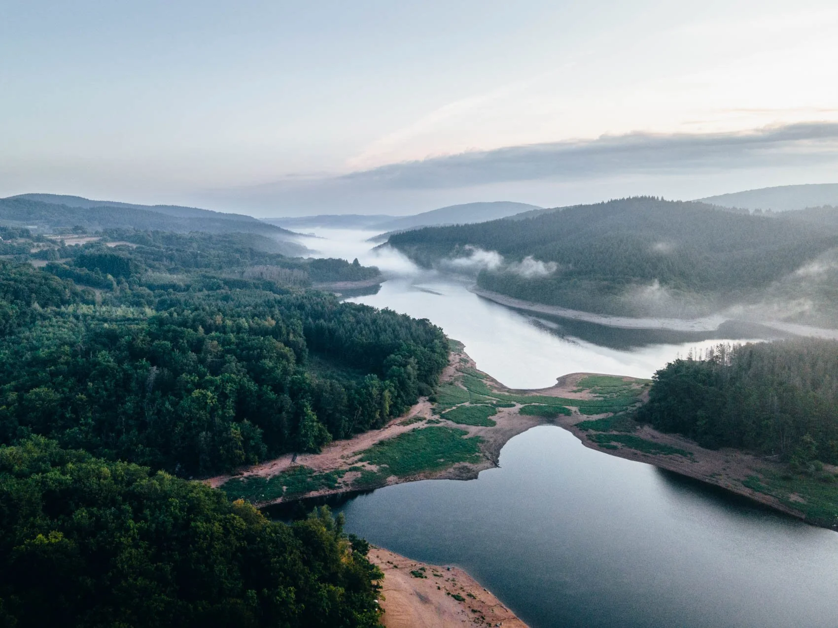 Vue aérienne d'une rivière serpentant à travers une vallée verdoyante, entourée de collines et de montagnes brumeuses au loin, par une matinée brumeuse.
