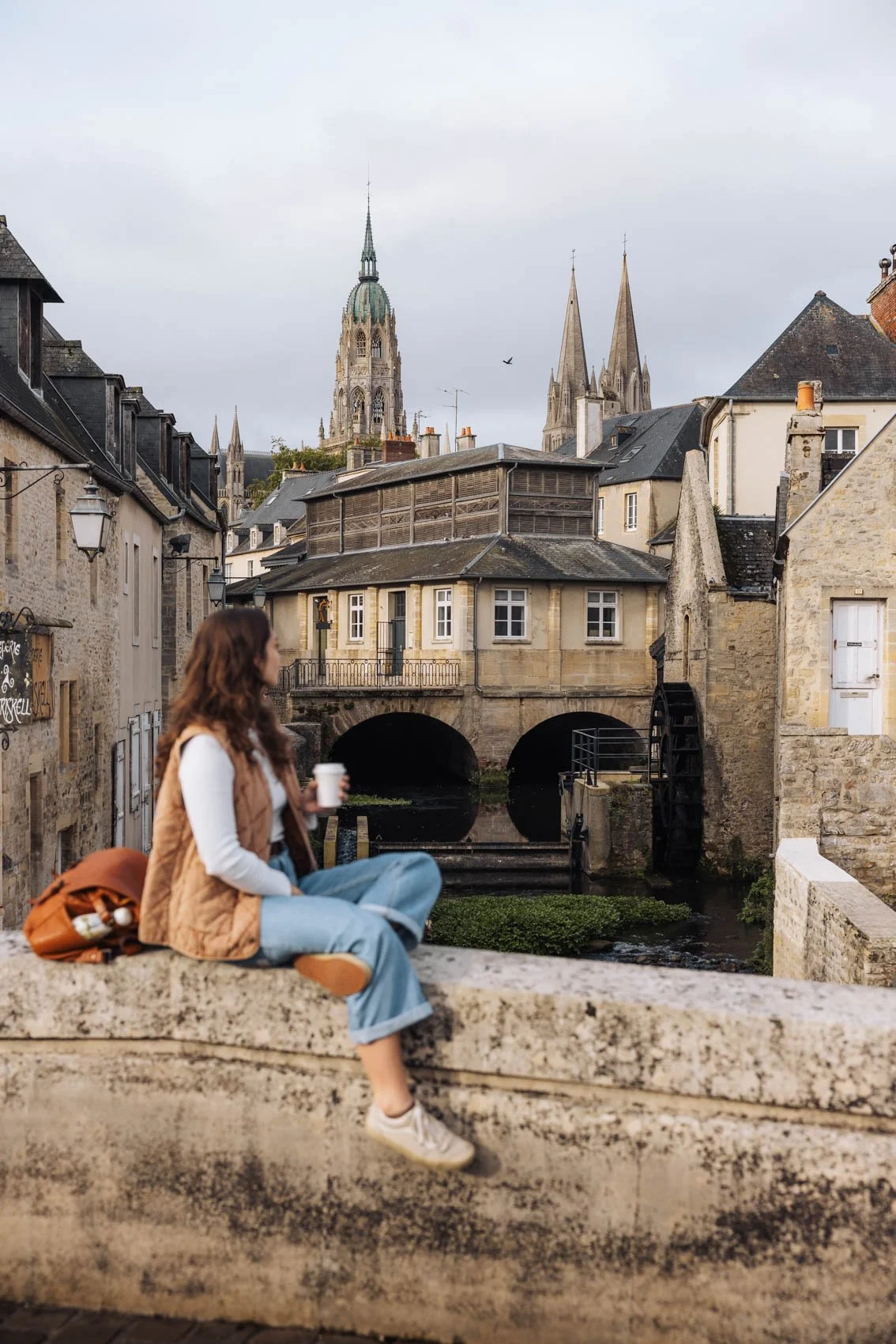 Une femme assise sur un mur en pierre, tenant une tasse, avec un village historique en arrière-plan, comprenant des bâtiments en pierre, une petite rivière avec un pont et des églises avec des tours.
