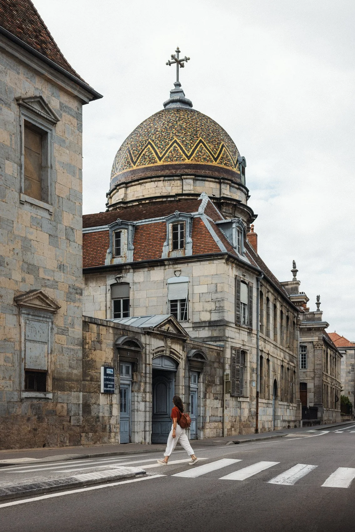 Une personne marche dans une rue pavée devant un vieux bâtiment en pierre avec un dôme coloré et une croix au sommet, sous un ciel nuageux.