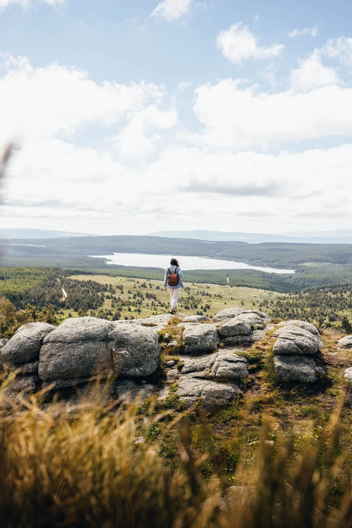 Une personne avec un sac à dos marche sur des rochers en haut d'une colline, dominant une vaste étendue de forêt et un lac, sous un ciel partiellement nuageux.