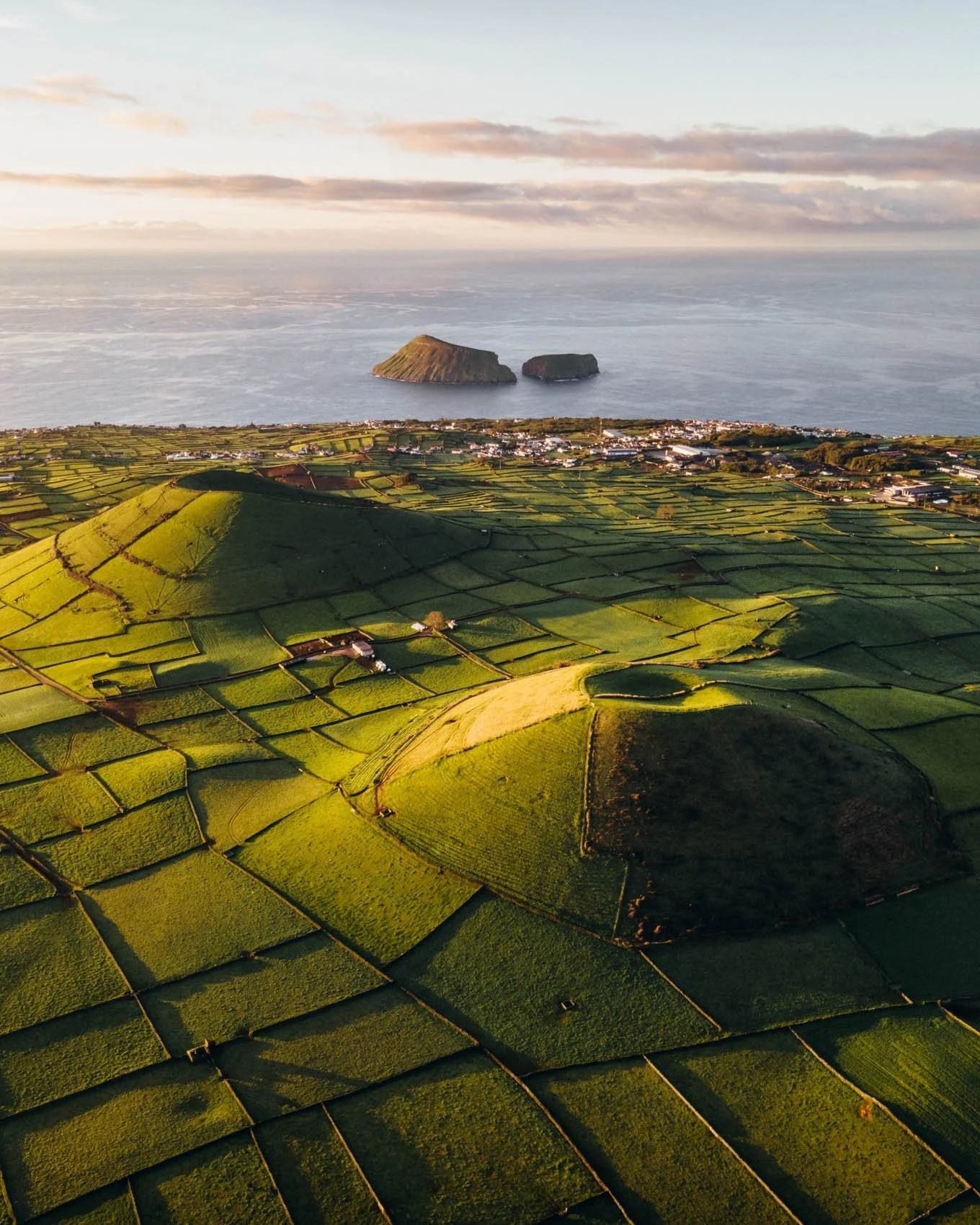 Photographie aérienne de champs agricoles verts en terrasses, bordée au loin par la mer avec deux îlots au large.