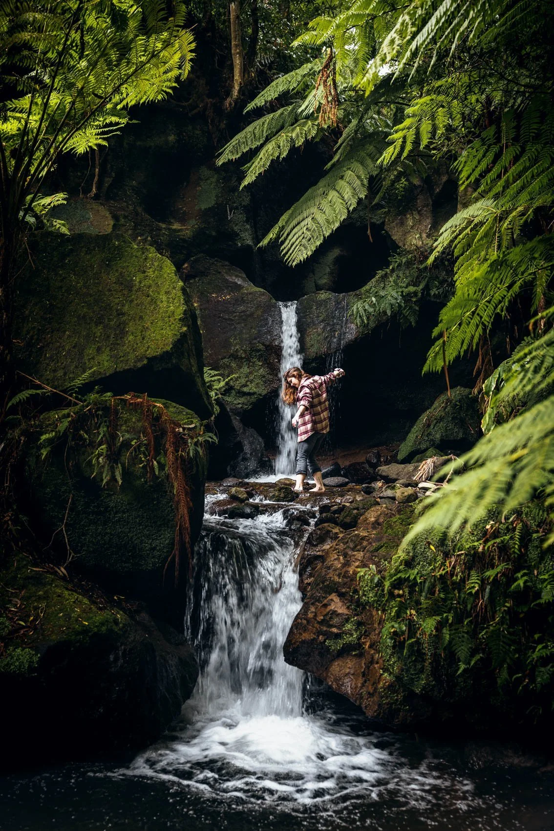 Une femme marche pieds nus dans une petite cascade entourée de végétation verdoyante et de rochers moussus dans une forêt dense.