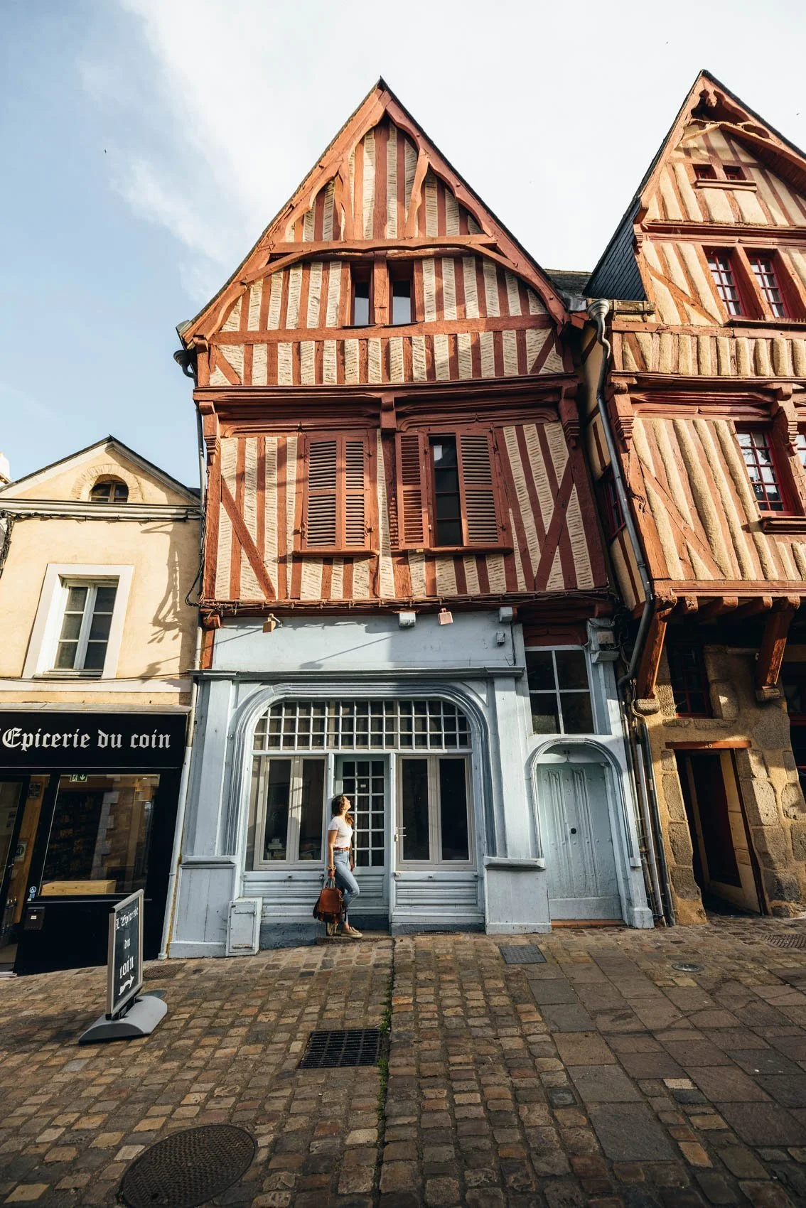 Une façade de bâtiment avec une partie en colombages en bois et une devanture blanche, un homme marche devant la boutique "Epicerie du coin" dans une rue pavée.