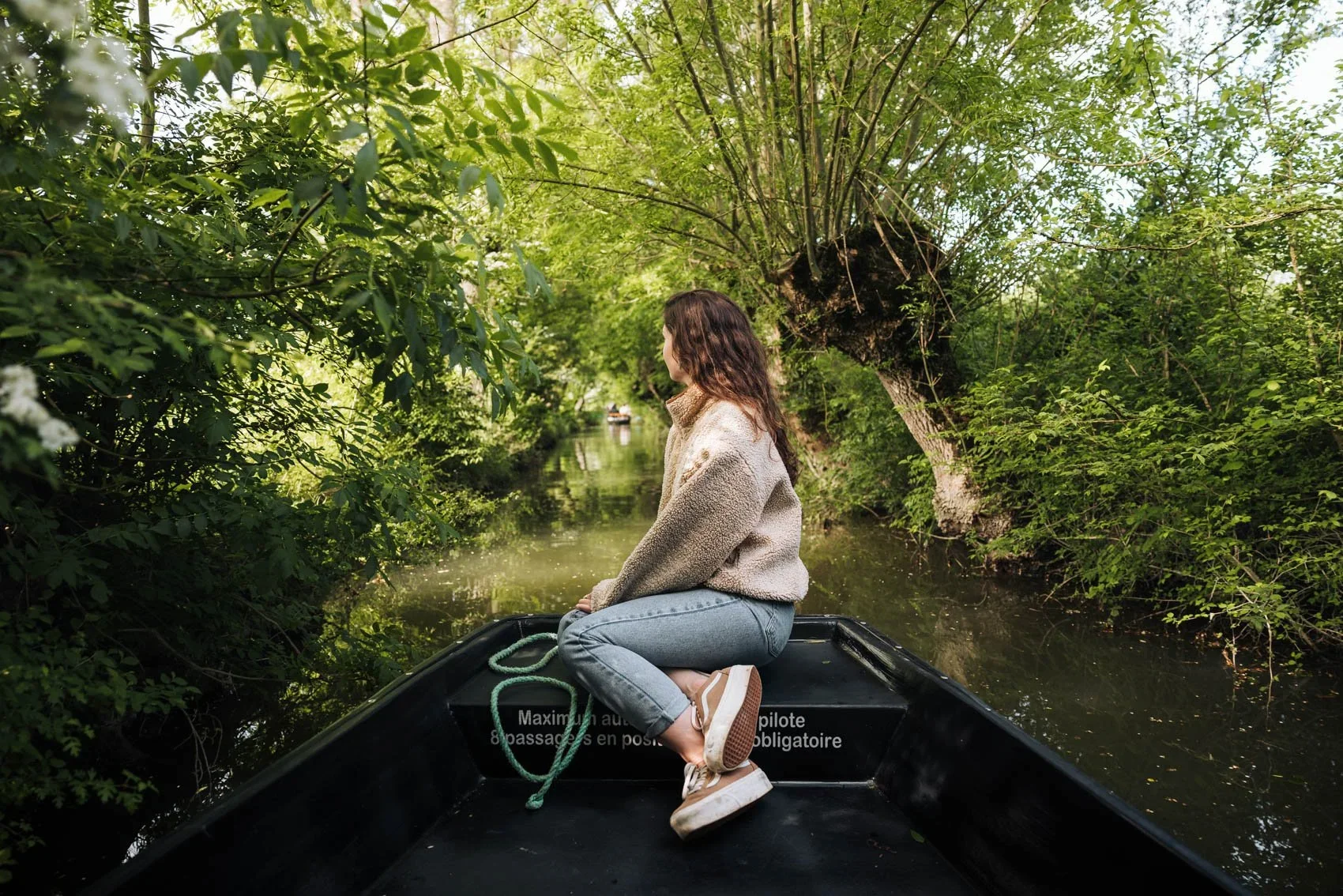 Une femme assise à l'avant d'un bateau naviguant sur une rivière entourée d'une végétation luxuriante, avec un arbre situé sur la rive. La scène semble calme et paisible, dans un environnement naturel verdoyant.